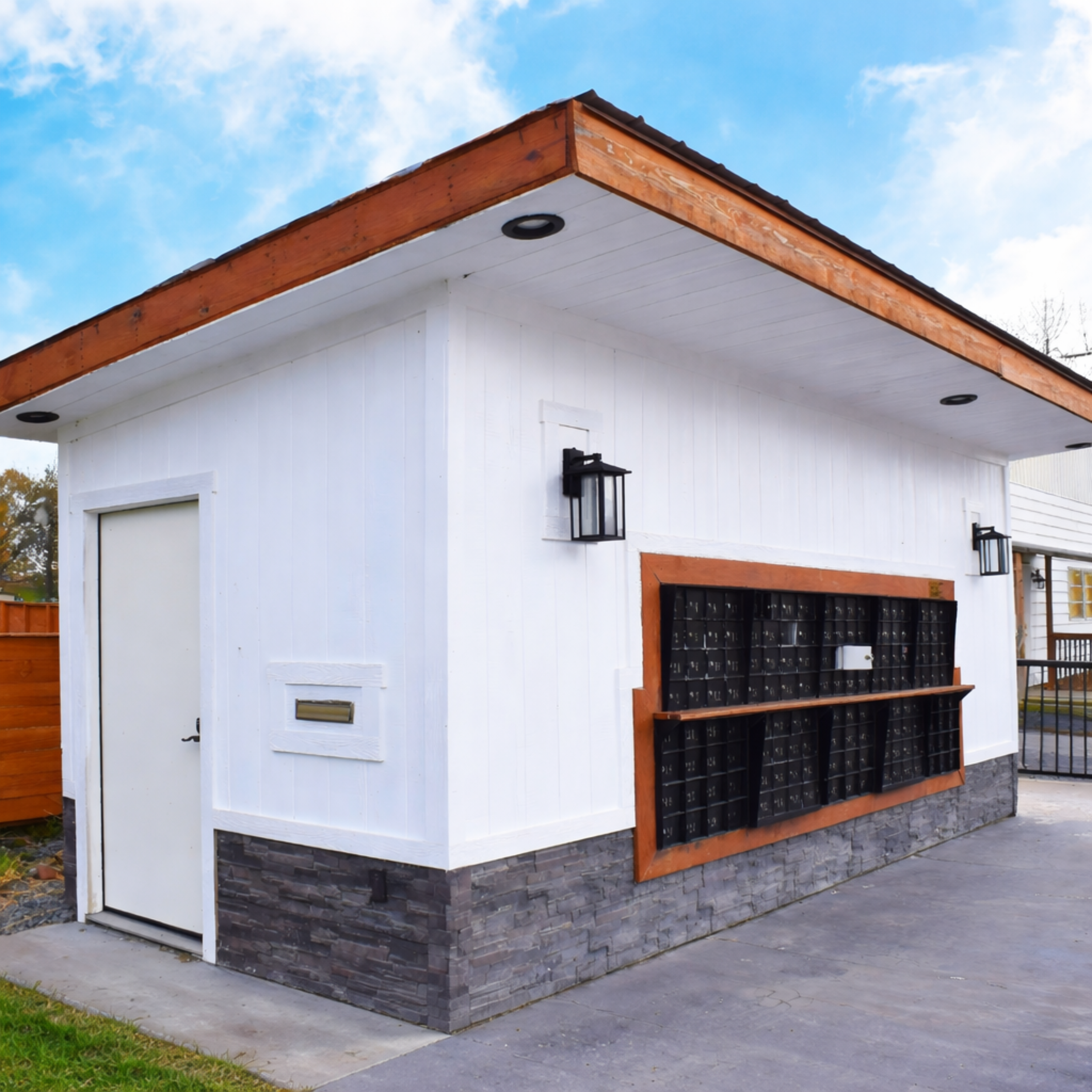 A small white building with a stone base, black mailboxes, and black outdoor lights. Wooden trim and a blue sky in the background.