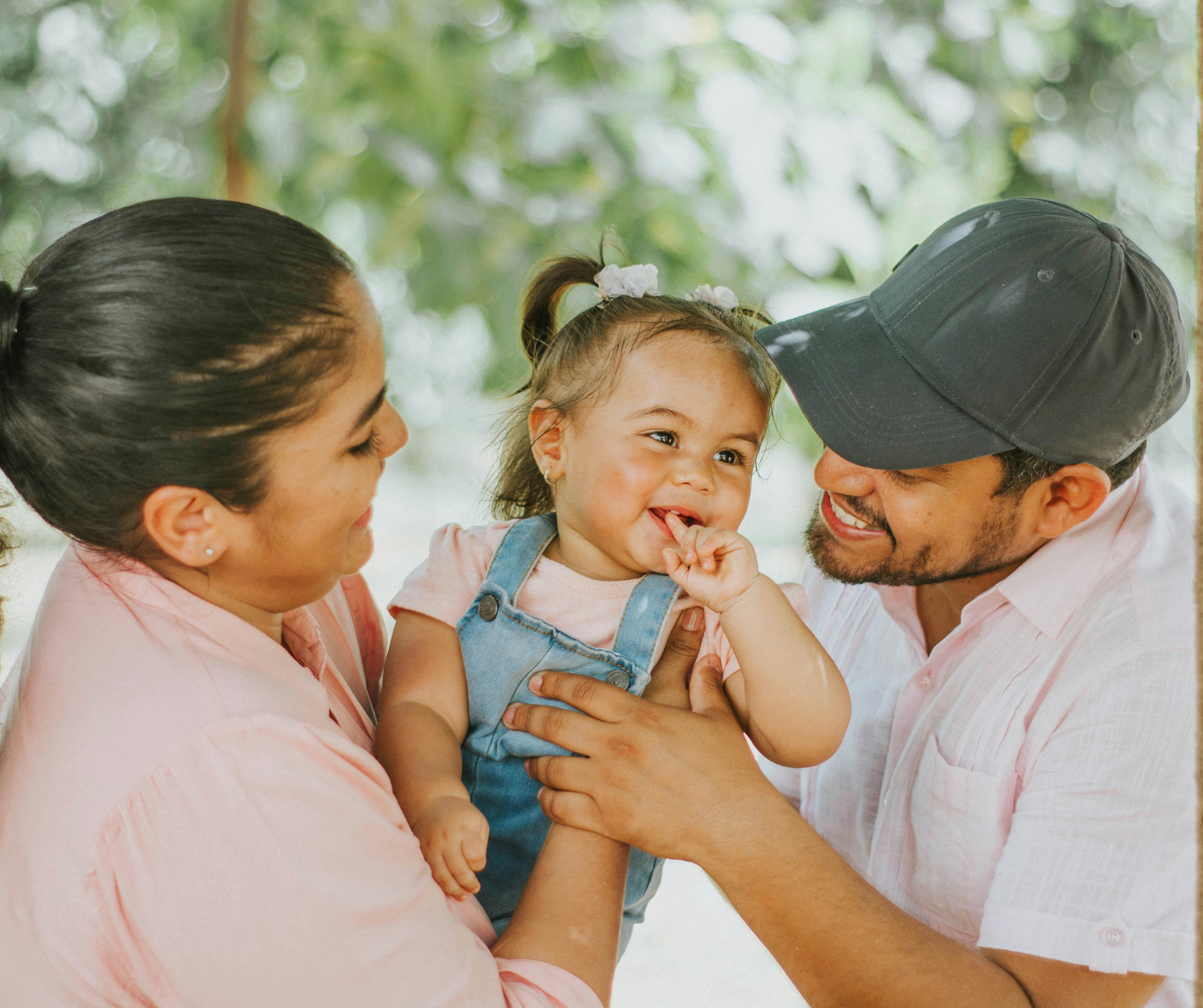 A family of three, a mother and father, holding their smiling young daughter outdoors surrounded by green foliage.