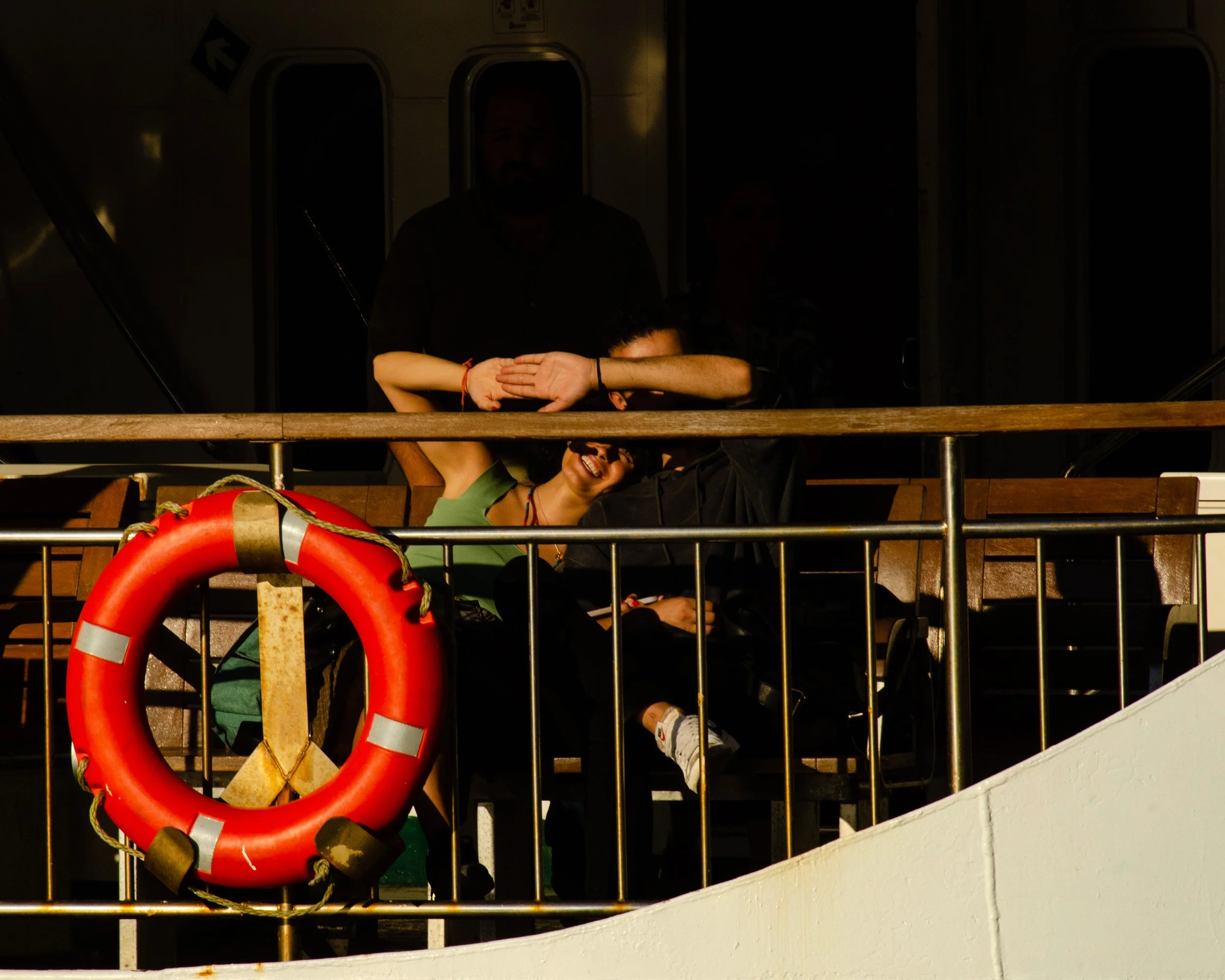 Pessoa sorrindo, sentada em um banco, com mãos atrás da cabeça, ao lado de uma pessoa deitada, dentro de um ambiente fechado com janelas, uma boia de vida vermelha pendurada na grade de proteção.