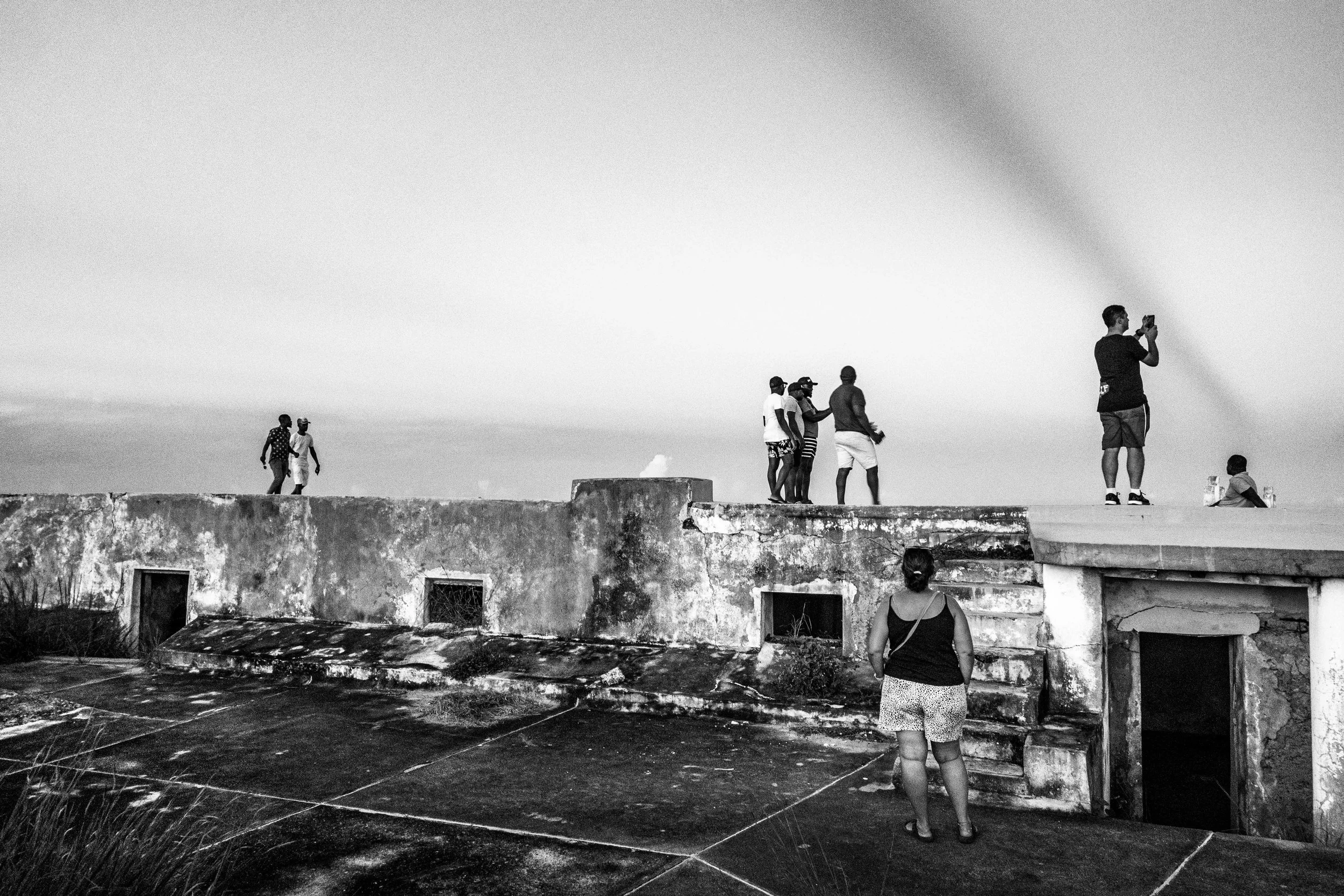 Grupo de pessoas em um terraço ao ar livre, alguns tirando fotos, outros conversando, com um prédio envelhecido ao fundo.