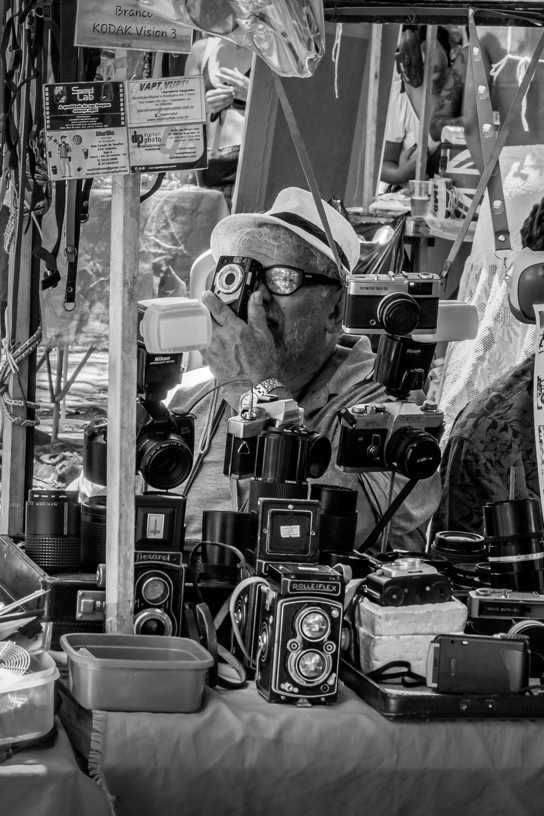 Homem mais velho usando óculos e chapéu, tirando uma foto com uma câmera fotográfica antiga em um mercado de rua que vende câmeras e acessórios fotográficos.