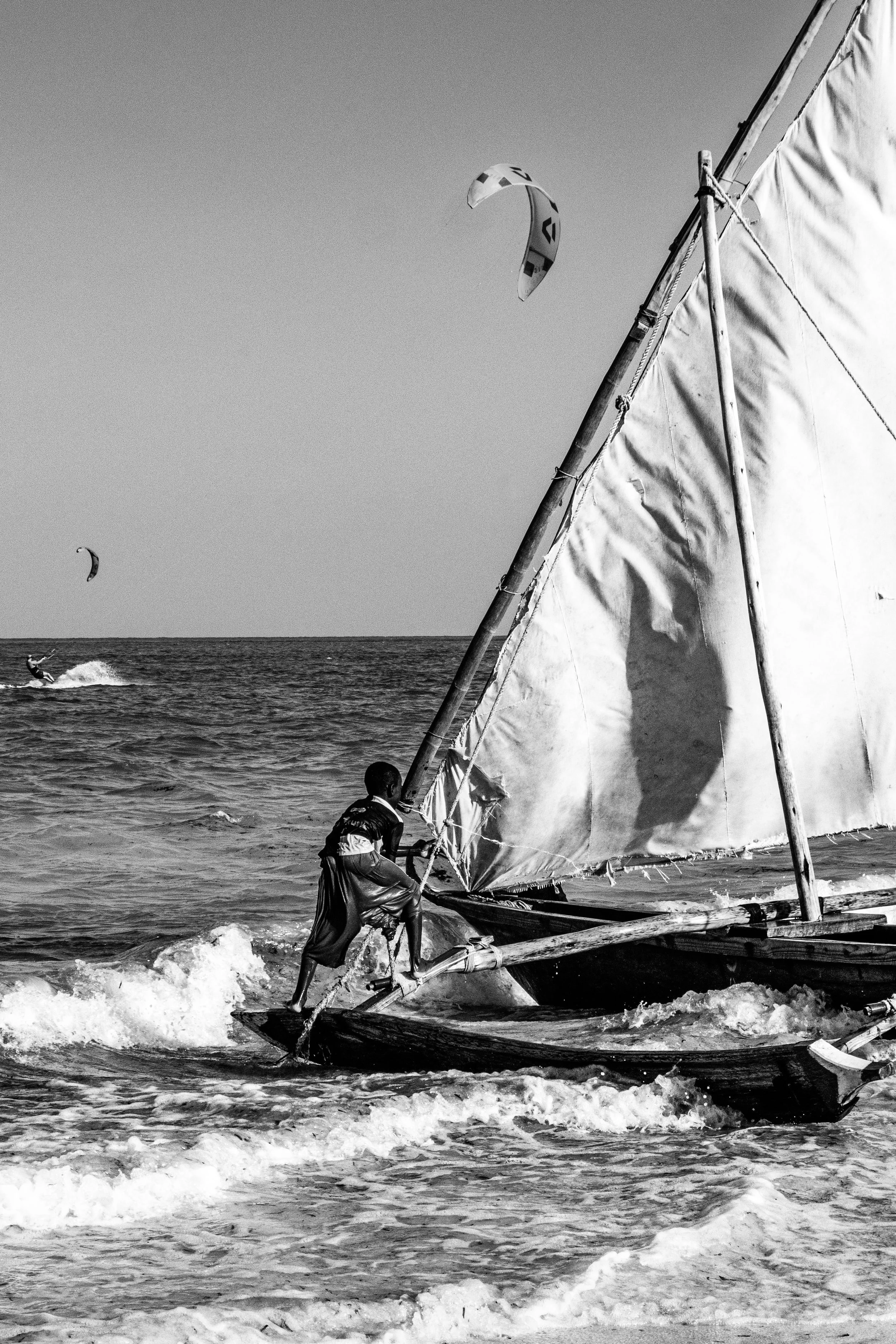 Jovem navegando em um barco a vela na praia, com duas pessoas praticando kitesurf ao fundo, tudo em preto e branco.