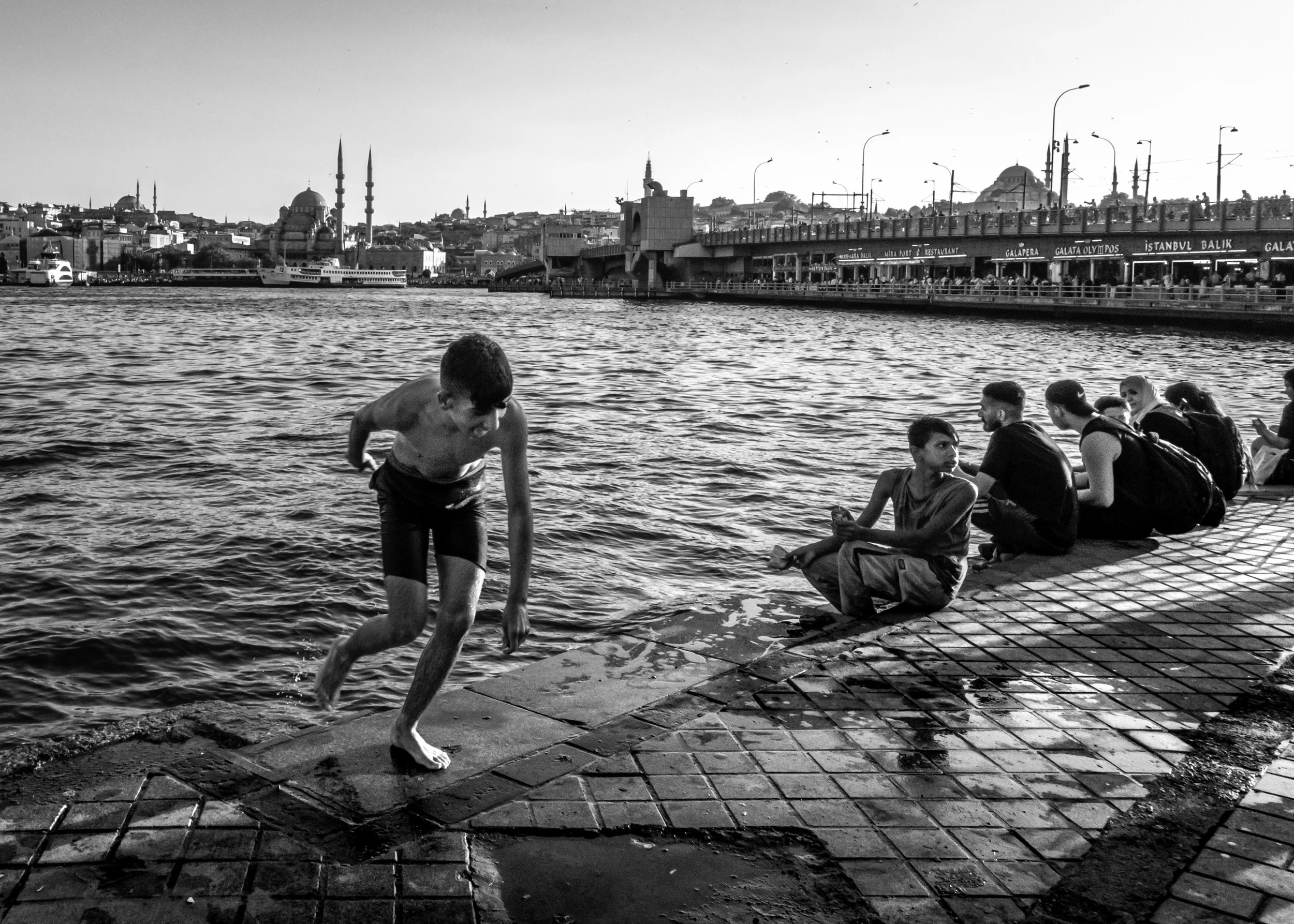 Criança pulando na água do rio, enquanto outras pessoas sentadas na margem observam, com uma cidade e uma ponte ao fundo, em uma cena urbana ao pôr do sol.