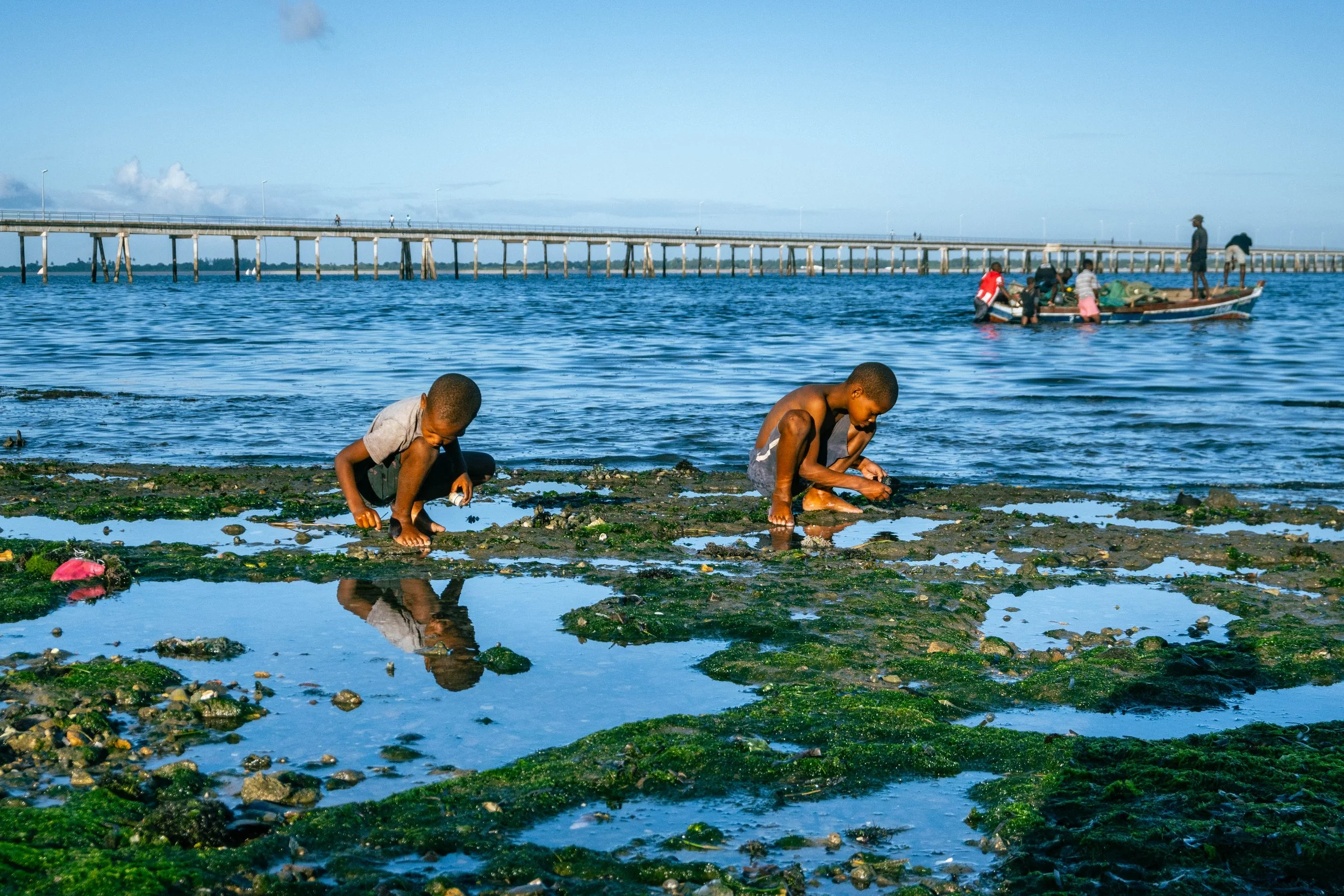 Dois meninos crianças brincando na água, agachados em uma praia de rio ou mar com pedras e algas, enquanto uma embarcação com pessoas está ao fundo e há um longo píer estendido sobre a água.