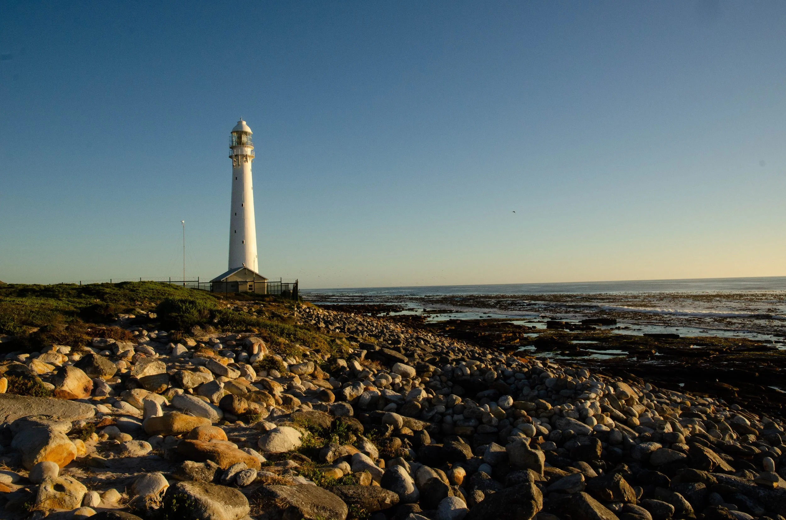Faro branco na costa rochosa ao pôr do sol, com céu limpo ao fundo.