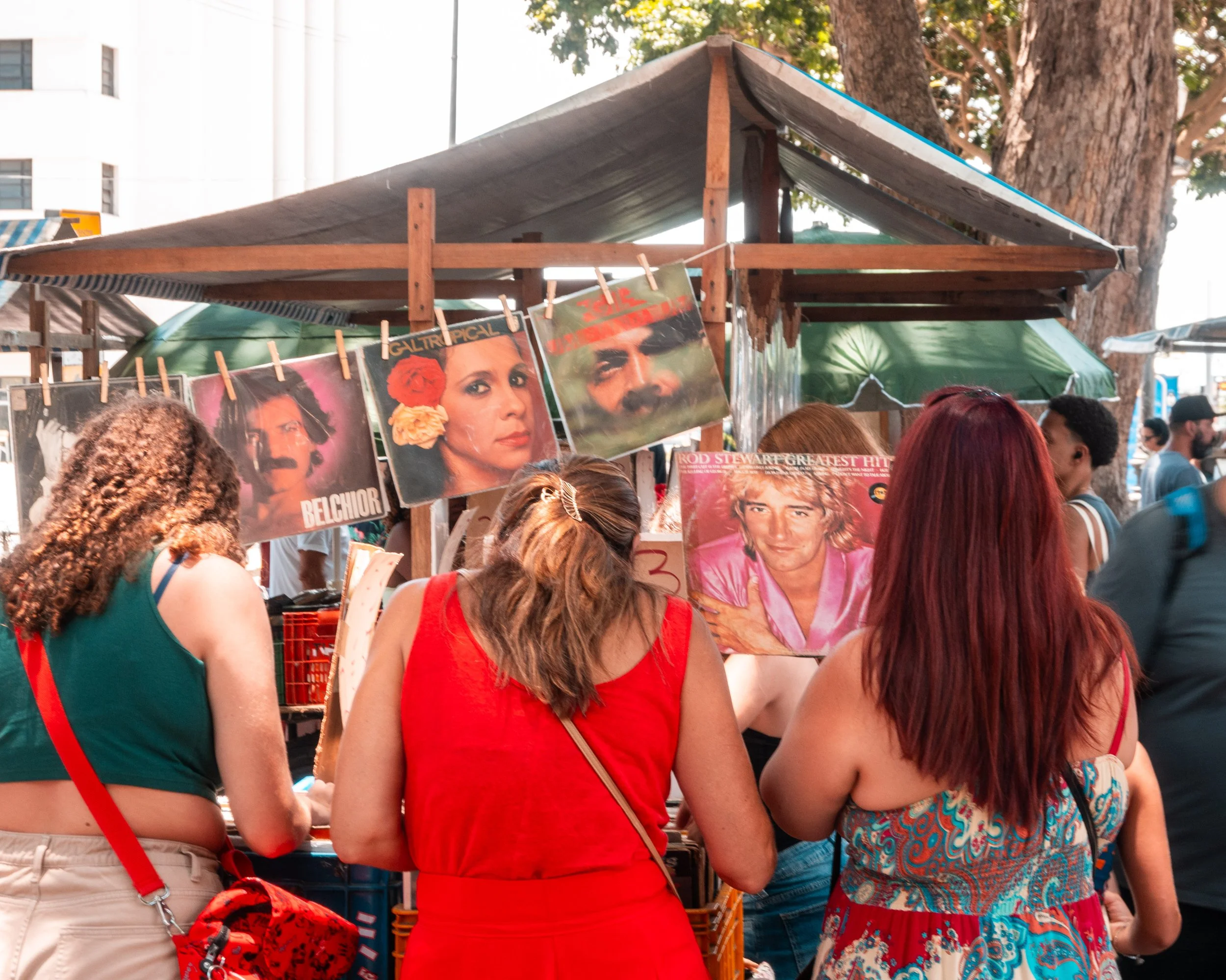 Vendedora de discos de vinil em feira ao ar livre, com várias pessoas ao redor, exibindo capas de álbuns famosos na estrutura de madeira.