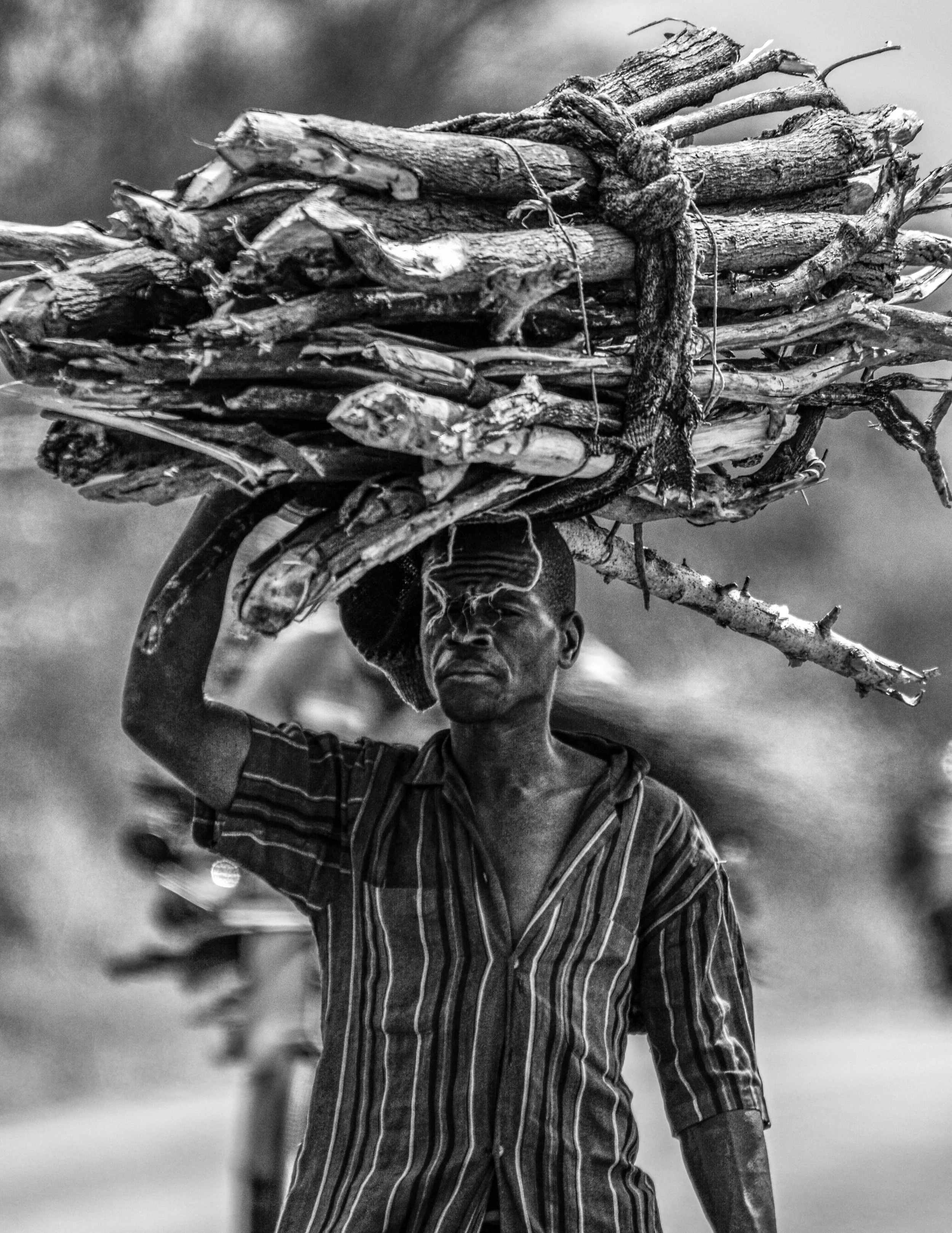 Homem carregando uma pilha de gravetos na cabeça, usando camisa de listras. Foto em preto e branco.