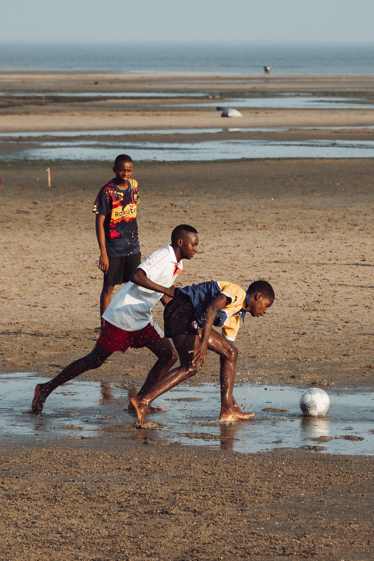 Três meninos jogando futebol na praia, com um deles encarando a bola, enquanto os outros dois estão atrás dele, um ao lado e outro no fundo.