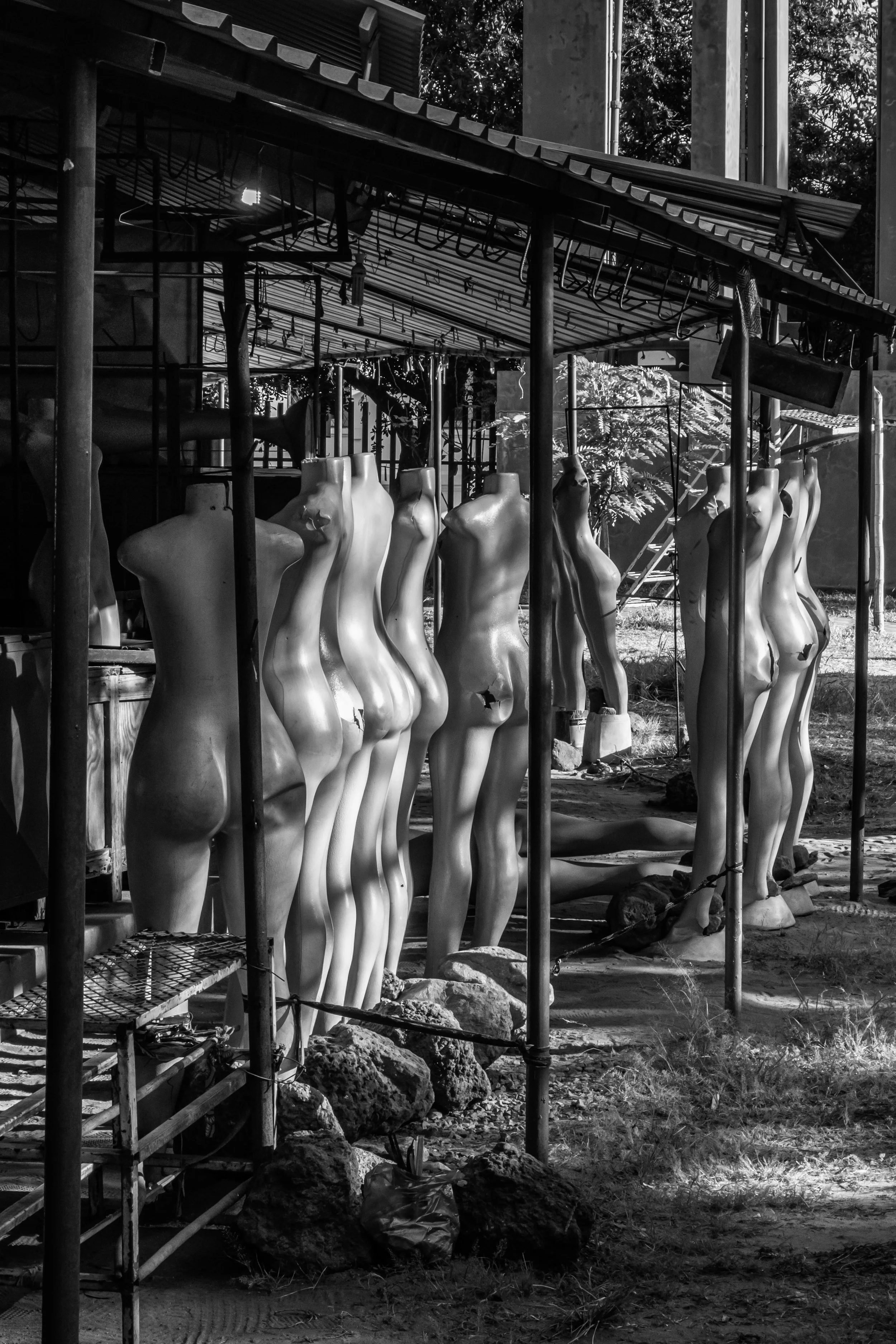 Esculturas de corpos femininos incompletos em uma área ao ar livre, cercadas por uma estrutura de metal e iluminação, em uma foto em preto e branco.