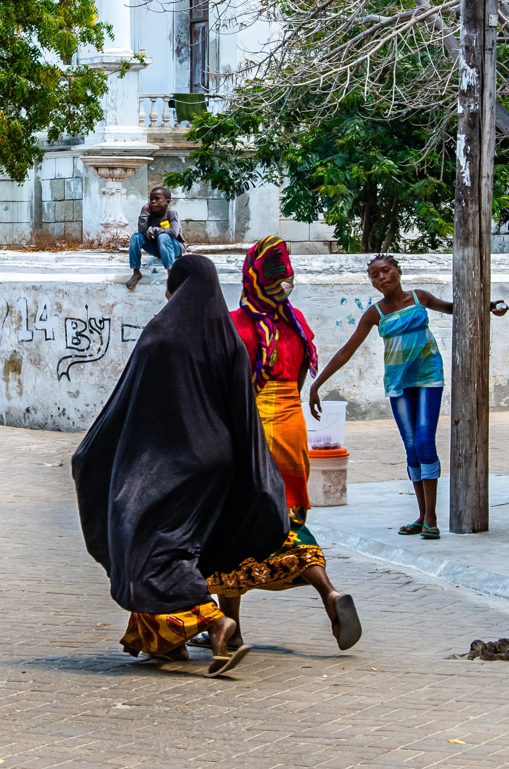 Quatro pessoas em uma rua, duas estão de frente para a câmera, uma vestida com roupas coloridas e uma coberta com um tecido preto, e uma criança está sentada em uma mureta ao fundo.