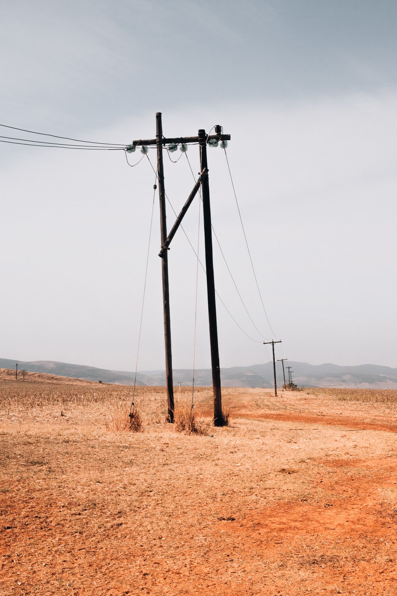 Postes de eletricidade em uma paisagem de campo árido com montanhas ao fundo e céu nublado.