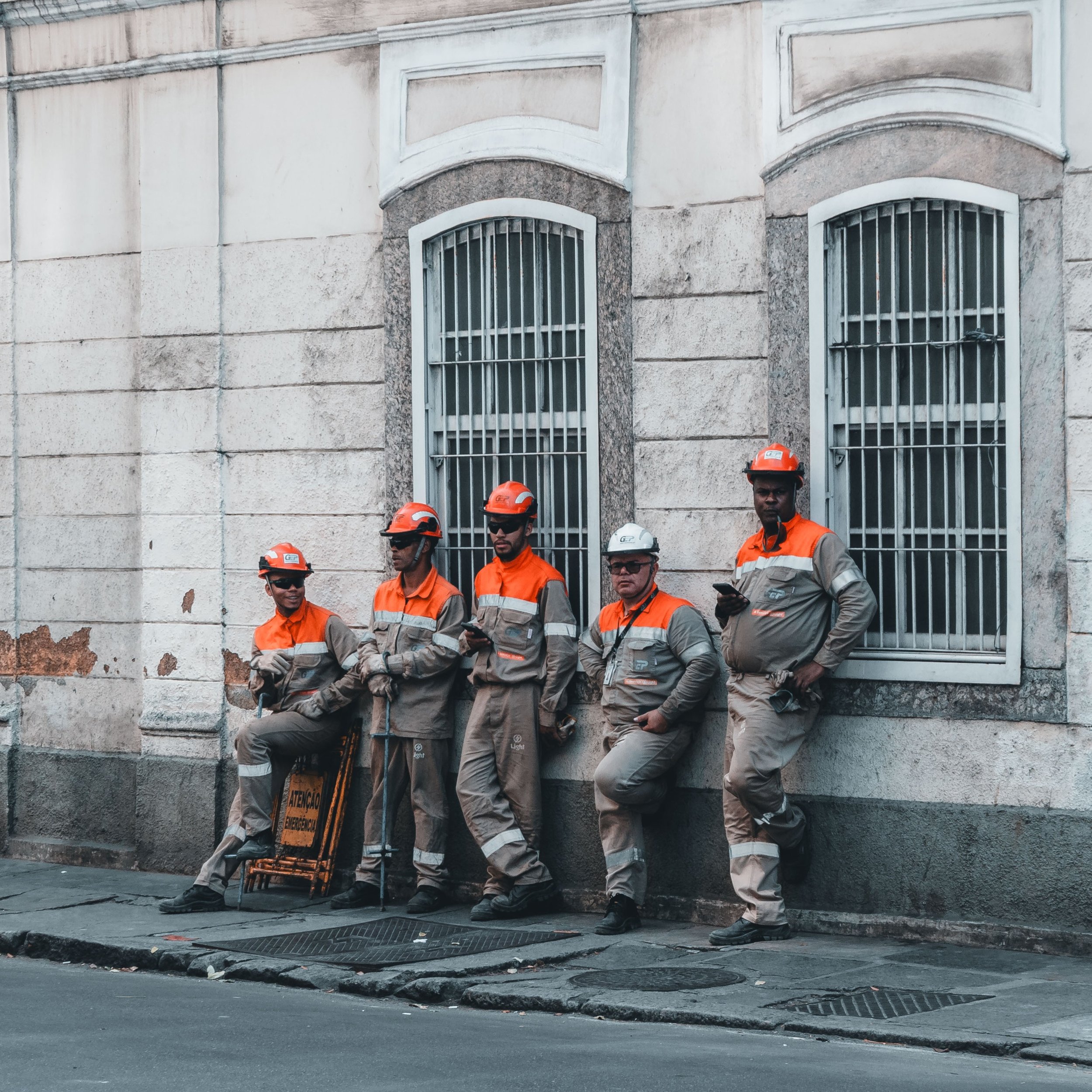 Cinco trabalhadores de uniforme laranja e cinza, com capacetes, descansando na calçada ao lado de uma parede de prédio com janelas de grades.
