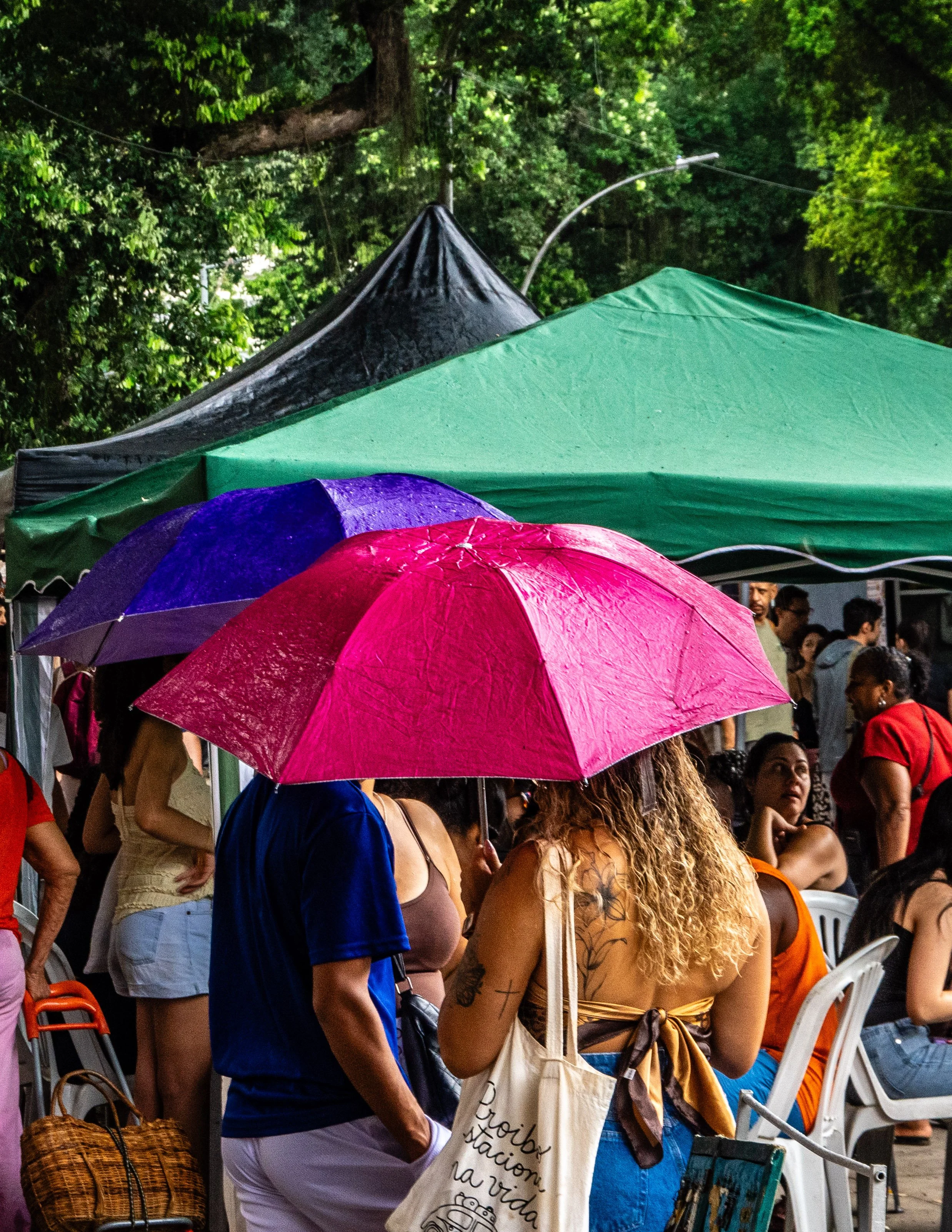 Mulheres com guarda-chuvas em evento ao ar livre sob chuva, com tendas e árvores ao fundo.