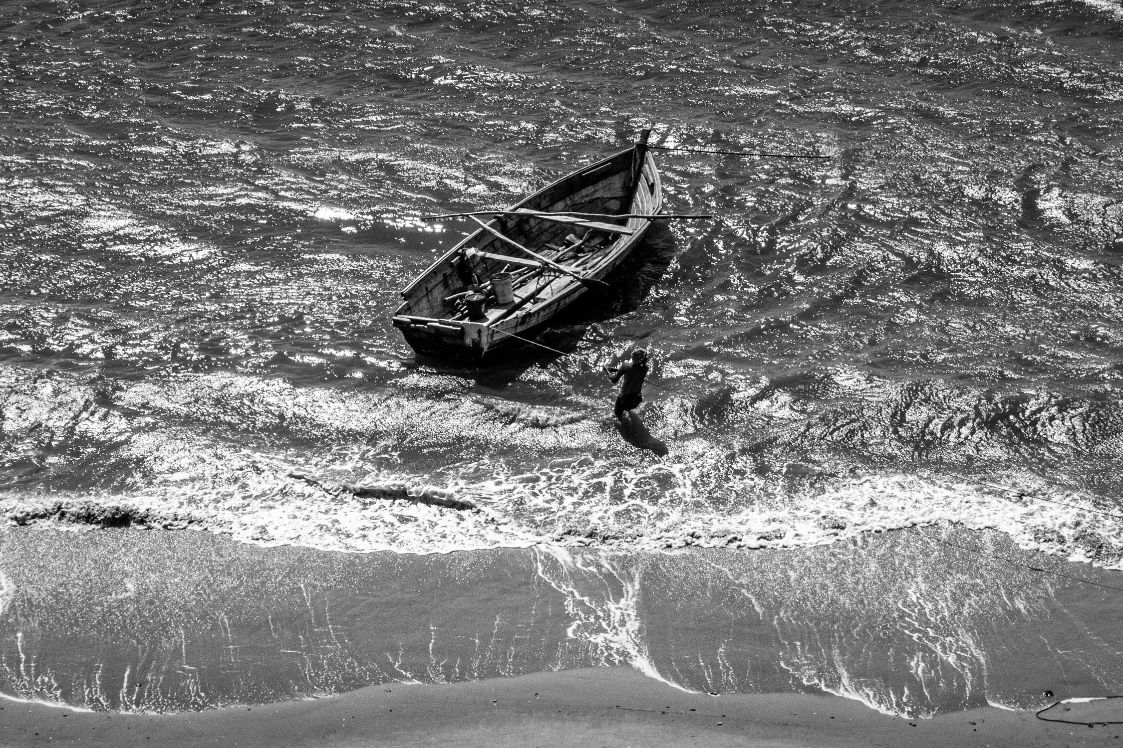 Homem surfando na água próxima a um barco de madeira na praia em uma imagem em preto e branco.