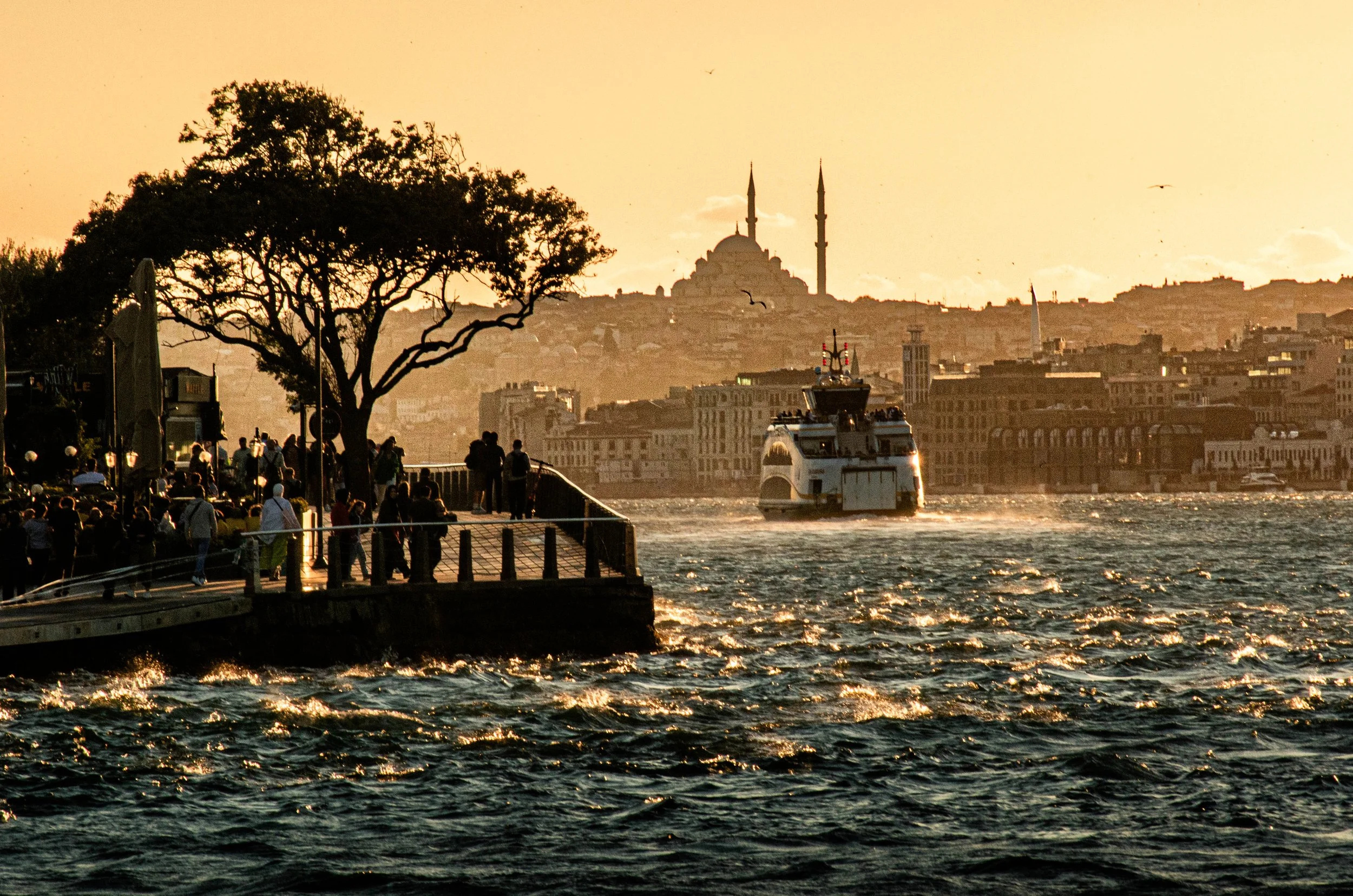 Cena ao pôr do sol na cidade de Istambul, com um cais ao lado do rio, pessoas caminhando, uma árvore grande, um barco navegando na água e a Silhueta da Mesquita de Sultan Ahmed ao fundo.