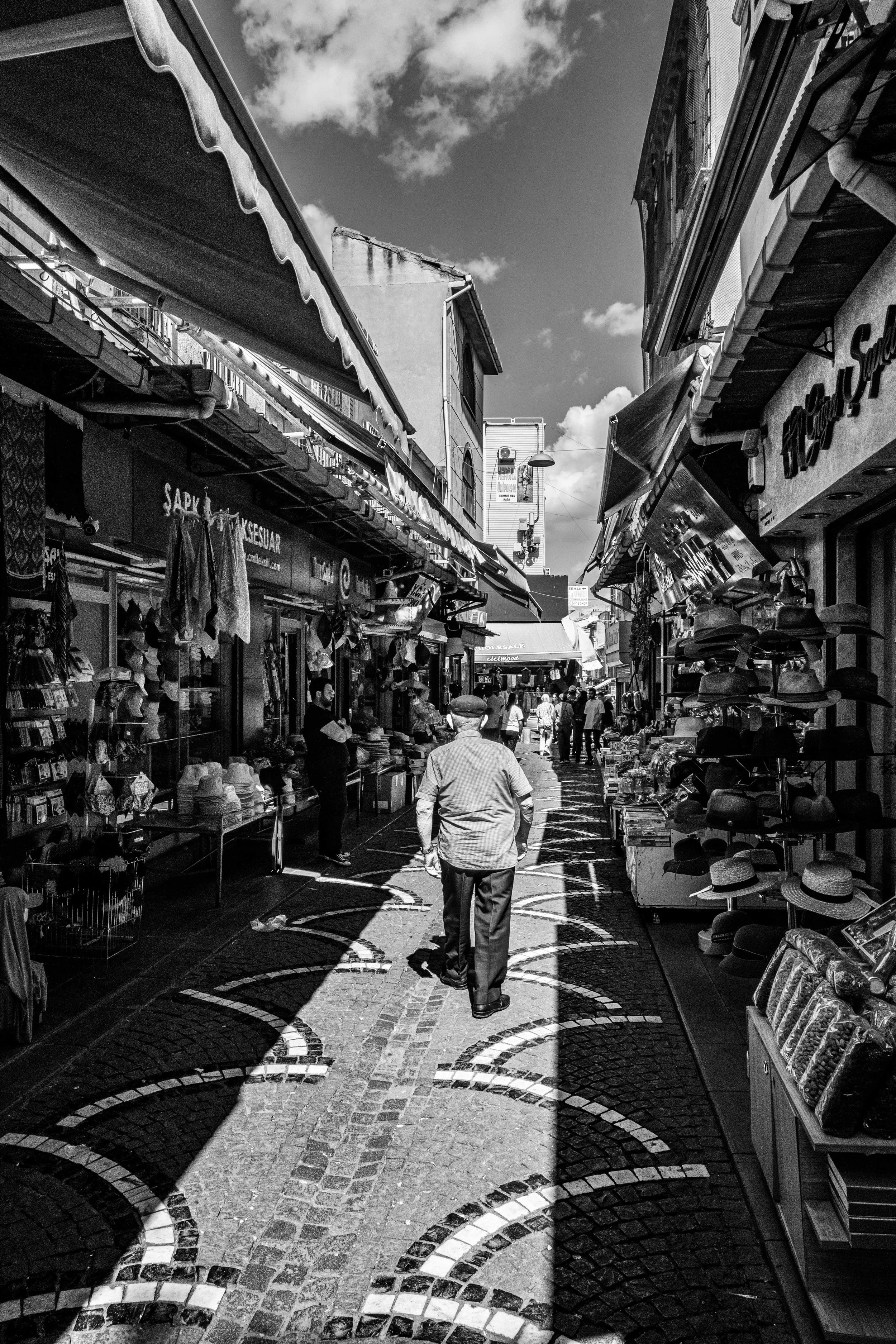 Rua de mercado ao ar livre com estandes de roupas, chapéus e souvenirs, pessoas caminhando sob guarda-sóis e fachadas de lojas, céu com nuvens.