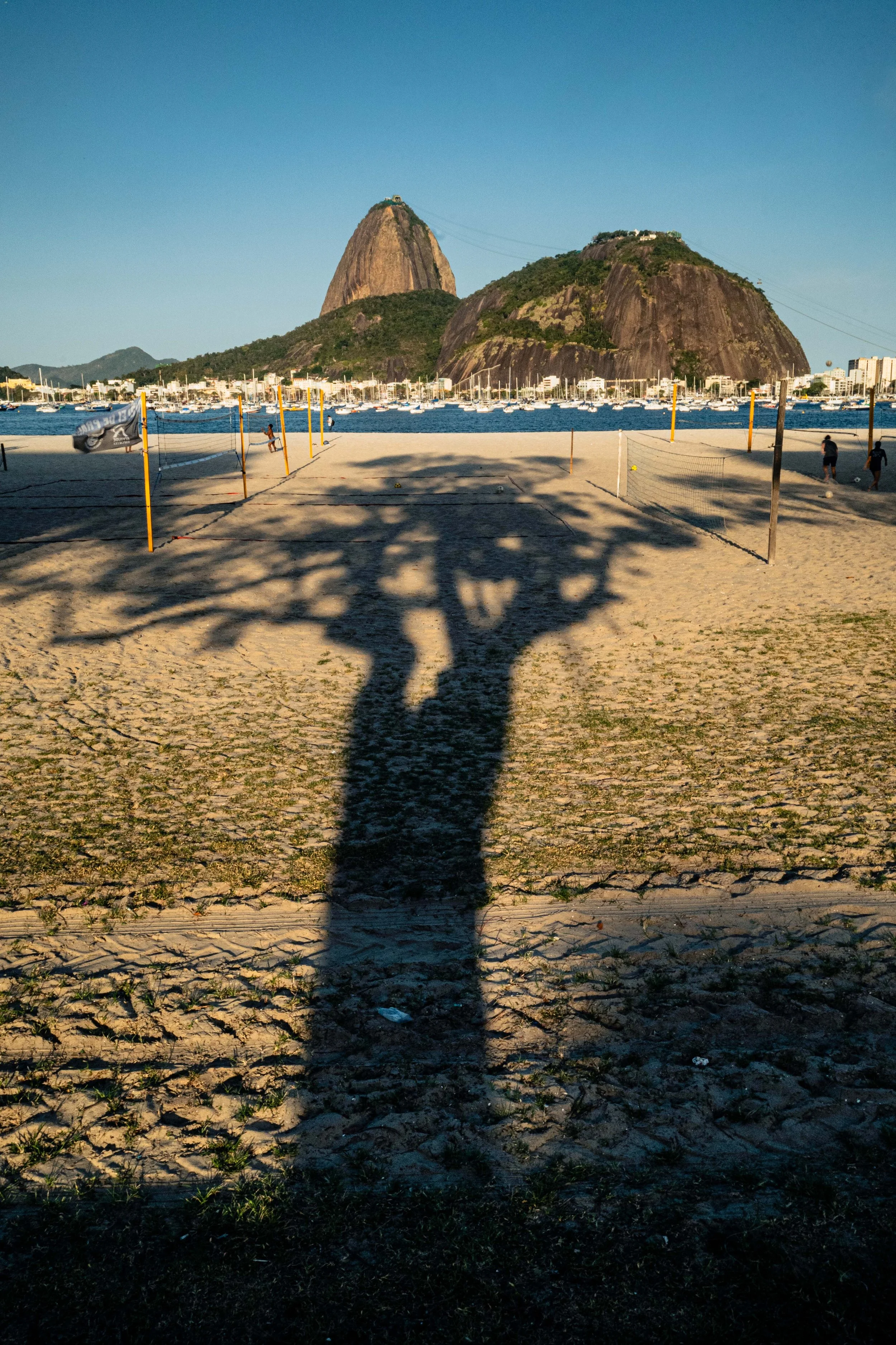 Sombra de uma árvore projetada na areia de uma praia, com quadras de vôlei, mar e montanhas ao fundo.