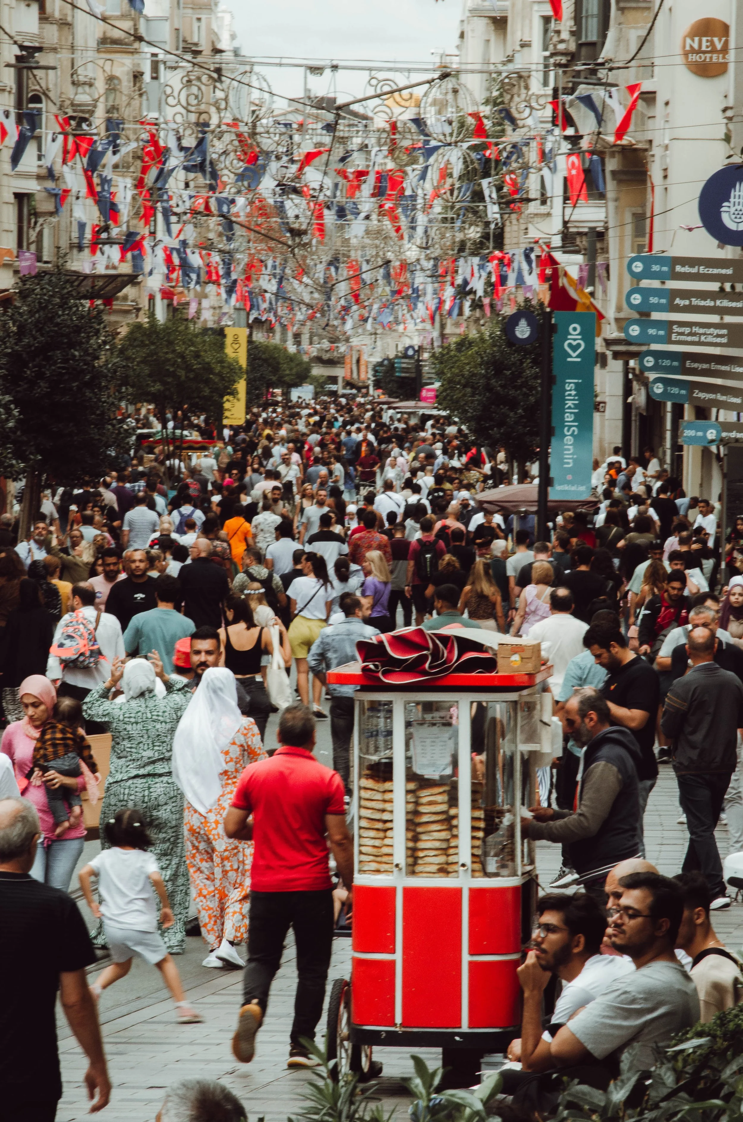 Rua movimentada em Istambul, Turquia, decorada com bandeiras e pessoas caminhando. Há um carrinho de comida com pratos empilhados na frente.
