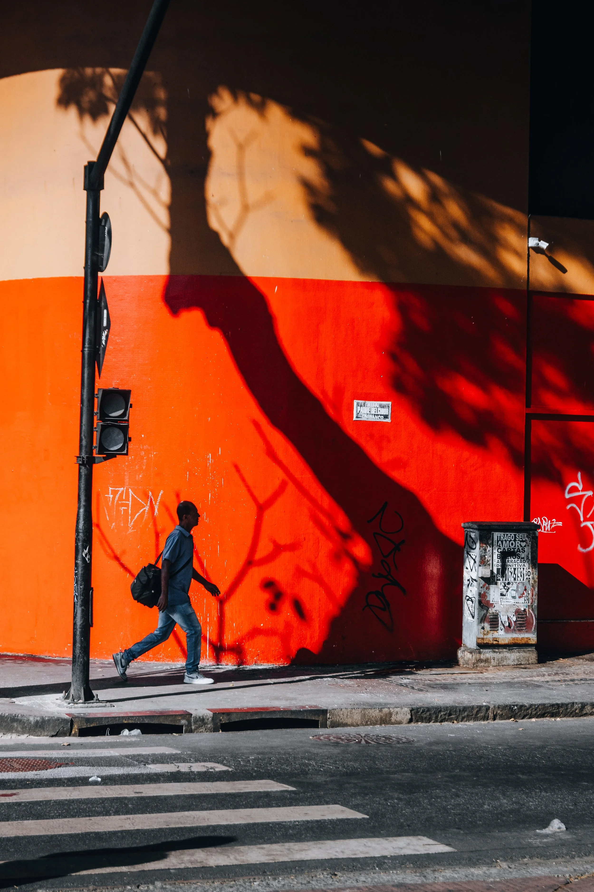 Homem caminhando na calçada ao lado de uma parede laranja com uma sombra de árvore projetada na parede, passando por um semáforo e uma caixa de mensagens de skate, com grafites na parede e papel de propaganda colado nela.
