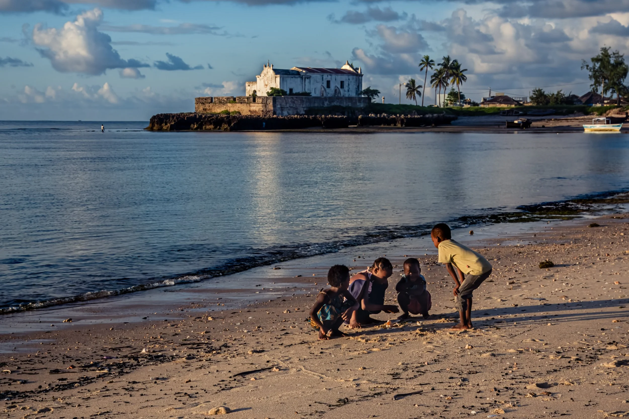 Crianças brincando na praia com castelos de areia ao entardecer, com uma fortaleza antiga ao fundo e palmeiras ao redor.