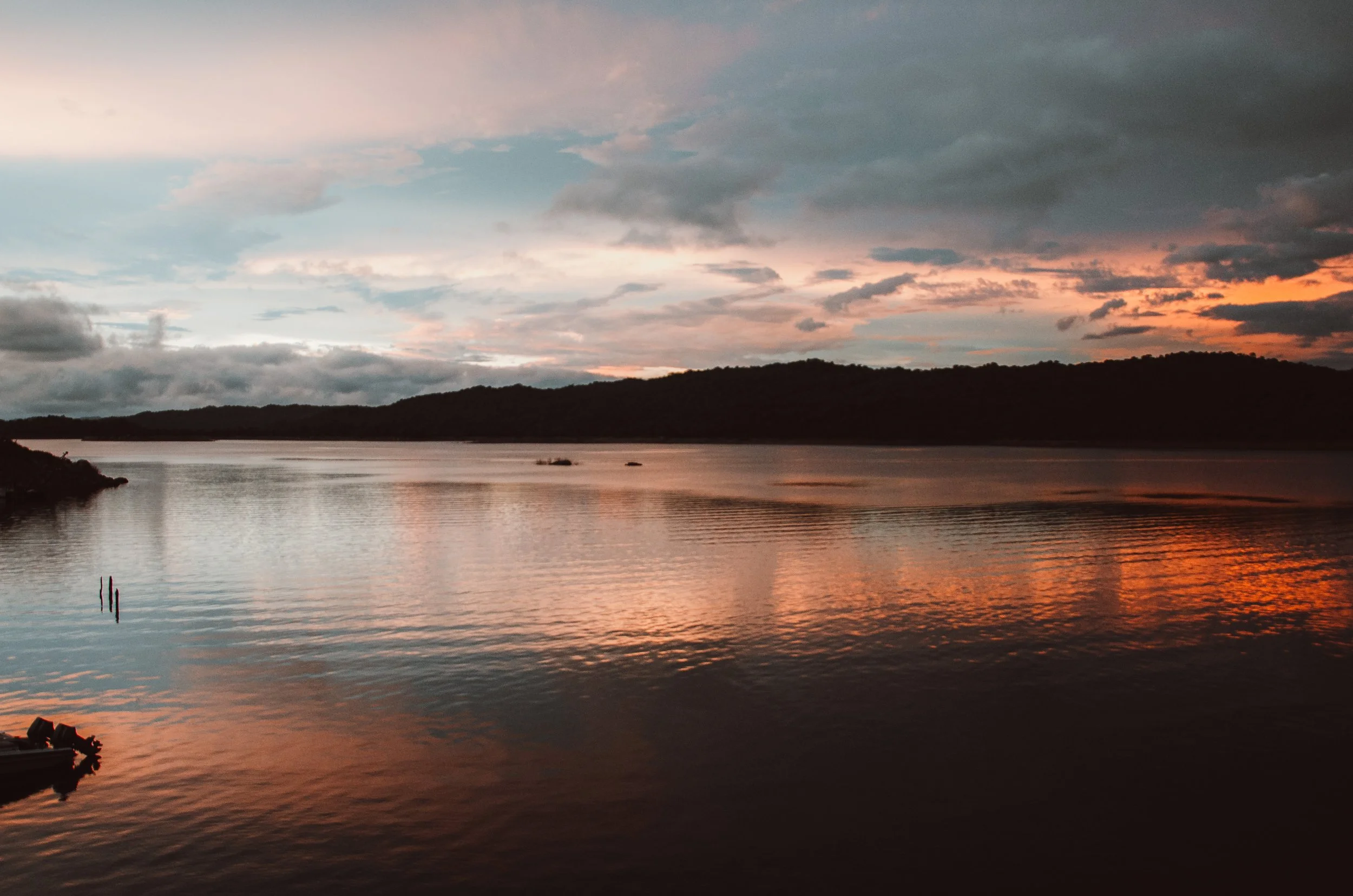 Céu ao pôr do sol com nuvens coloridas refletidas em um rio tranquilo, com uma margem de terra com vegetação ao fundo.