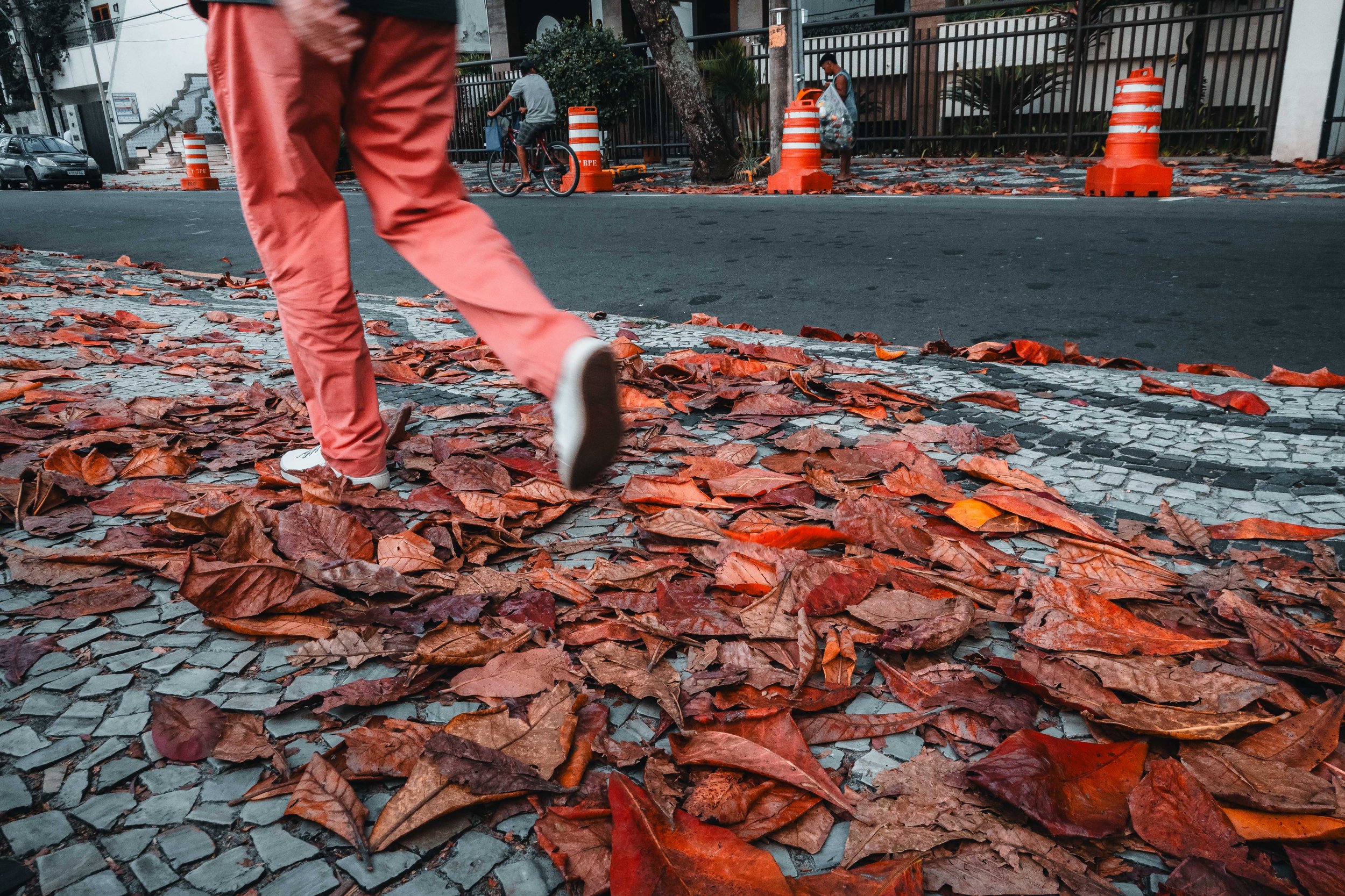 Pessoa caminhando sobre folhas secas na calçada de pedras em uma rua urbana com cones de trânsito ao fundo.