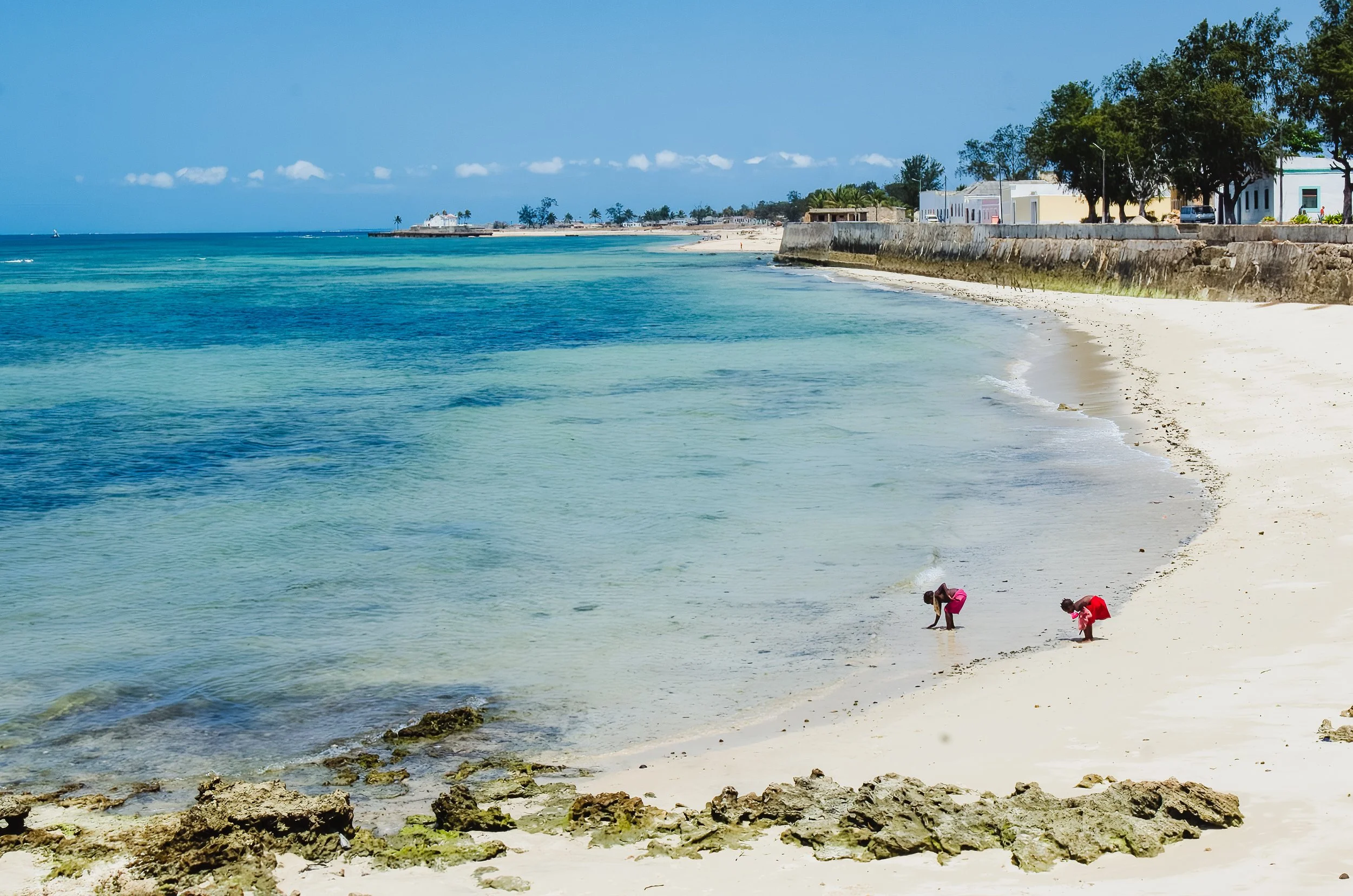Praia de areia branca com duas crianças brincando perto do mar, águas azuis e céu claro, com árvores e edifícios ao fundo.