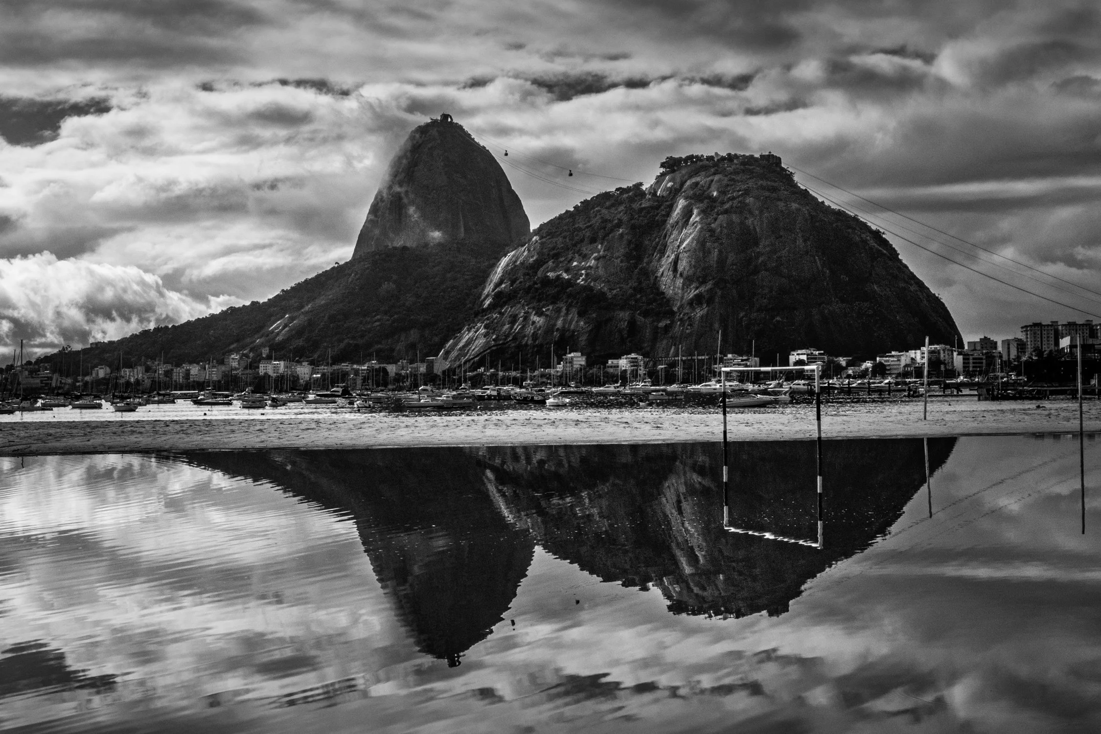 Paisagem urbana com duas montanhas grandes e um lago refletindo suas silhuetas, com barcos e prédios ao fundo, sob céu parcialmente nublado, em preto e branco.