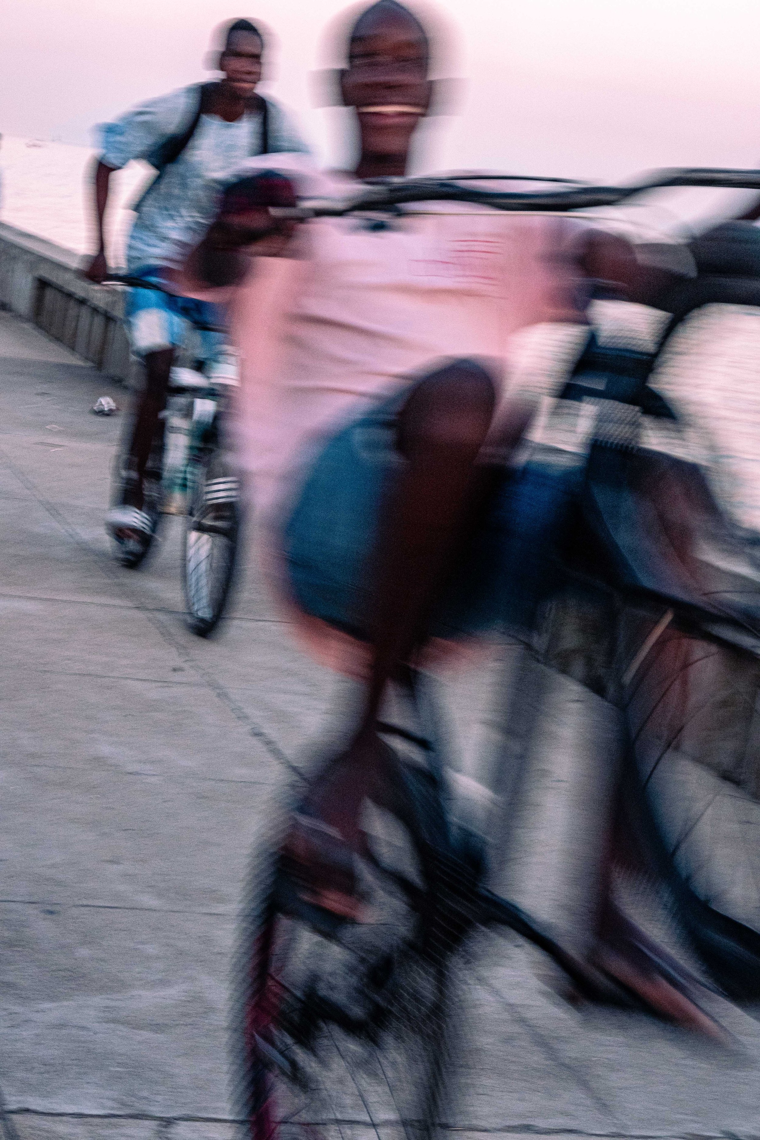 Imagem de duas pessoas andando de bicicleta na orla, uma delas sorrindo, com o mar ao fundo ao entardecer.