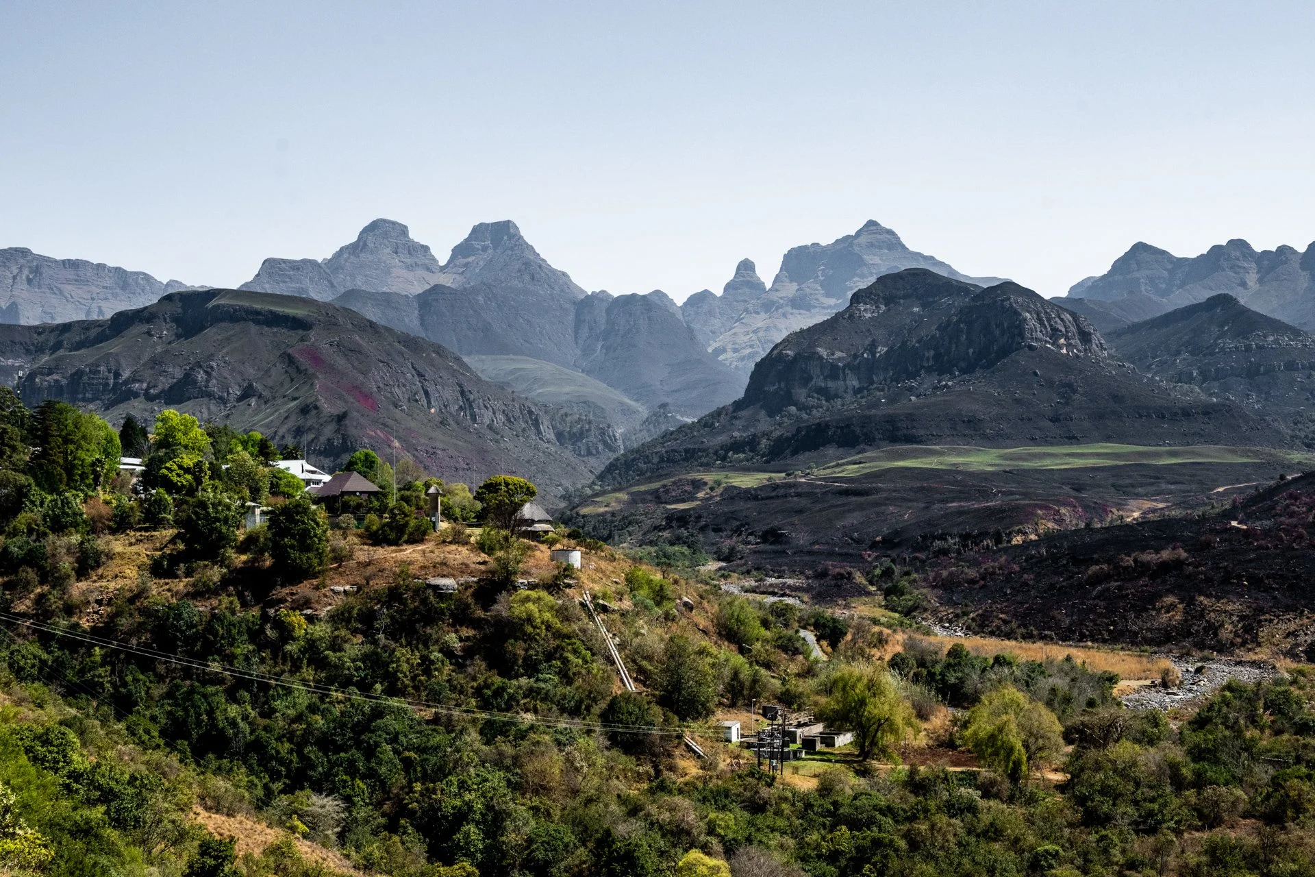 Paisagem de montanhas com vegetação verde no vale e casas ao longo da encosta, ao fundo montanhas rochosas sob céu claro.