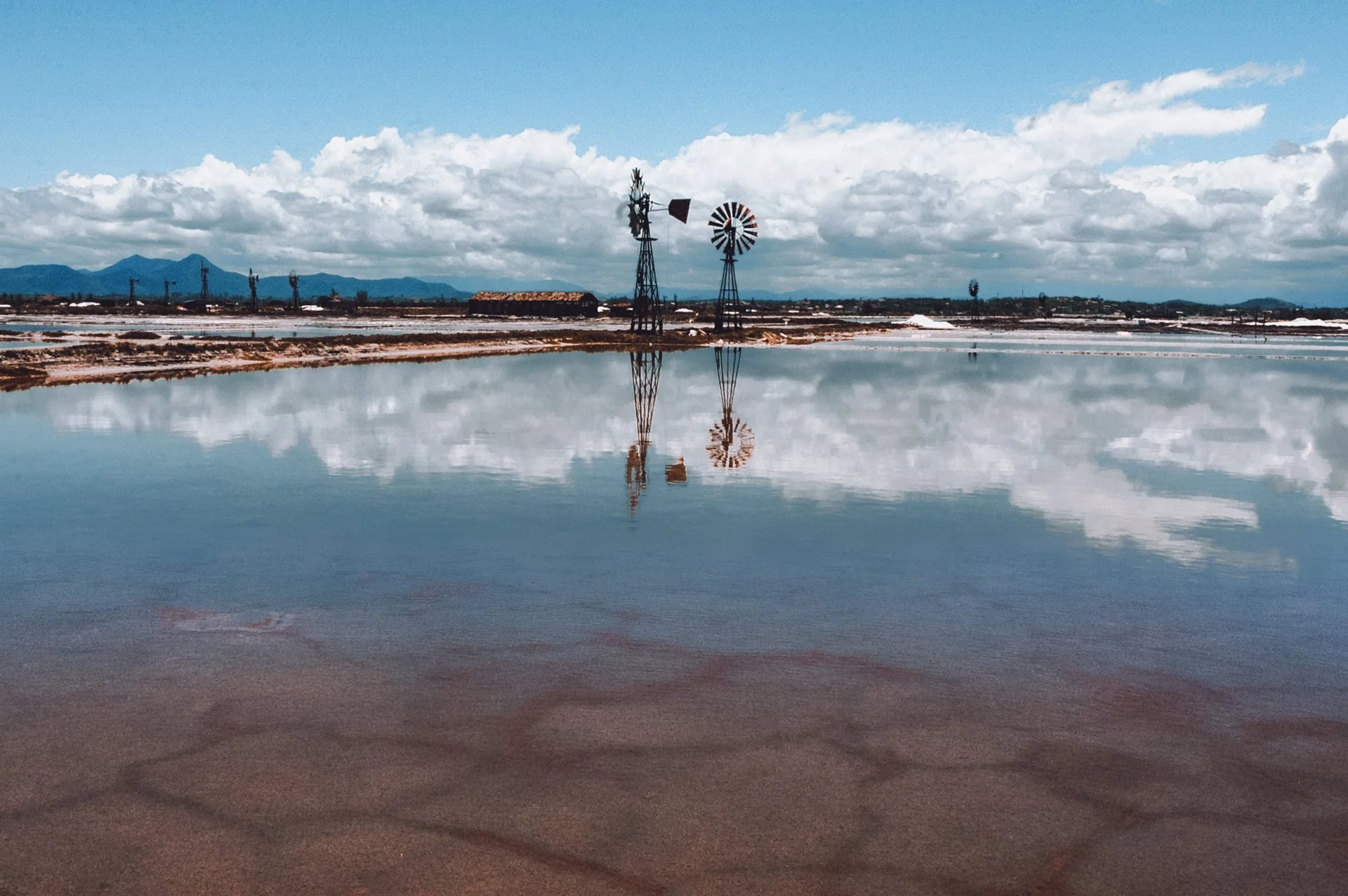 Reflexão de dois moinhos de vento de metal em uma lagoa sob céu com nuvens.
