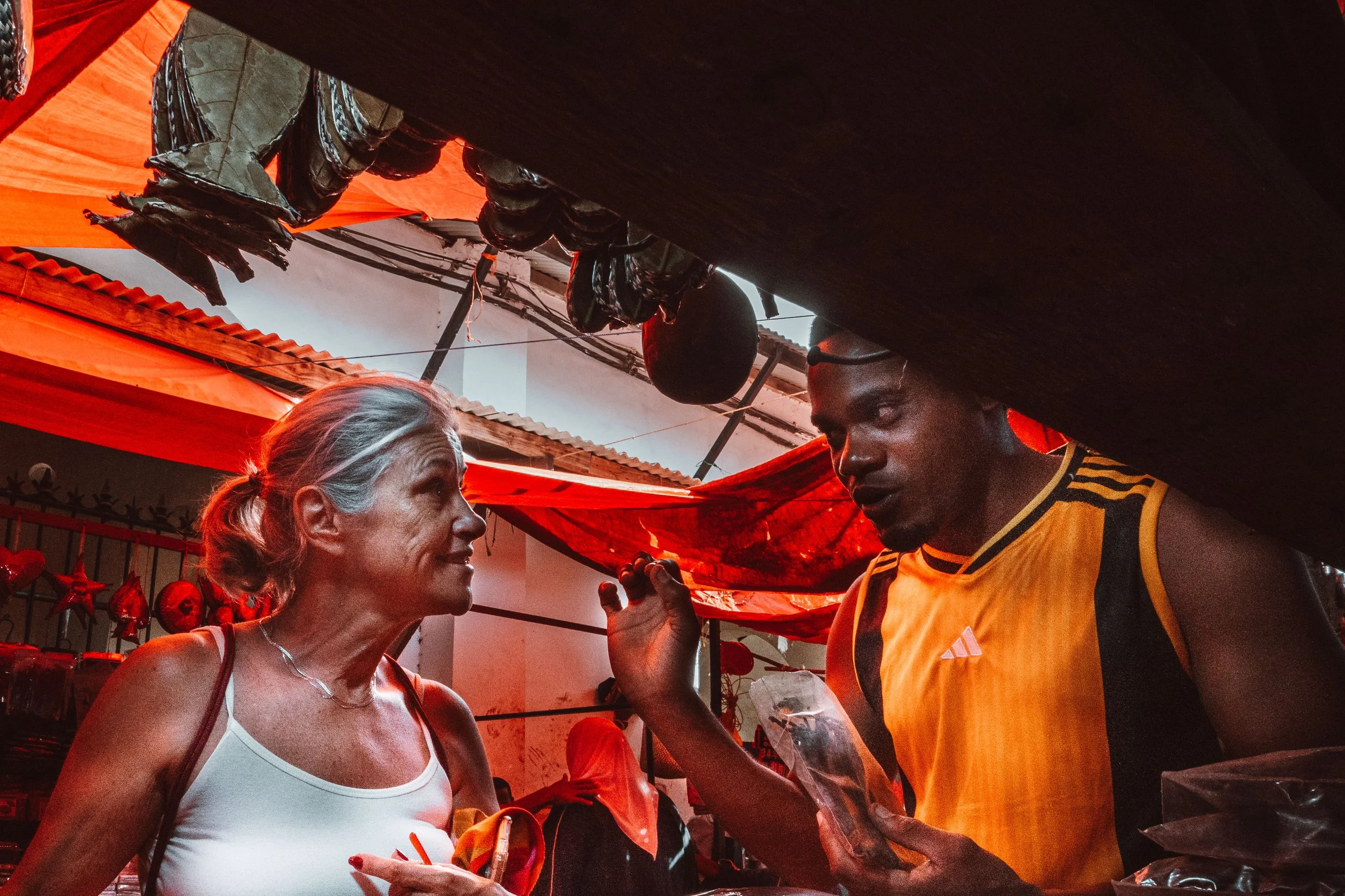 Mulher idosa conversando com jovem vendedor em feira com barracas cobertas por lonas vermelhas.