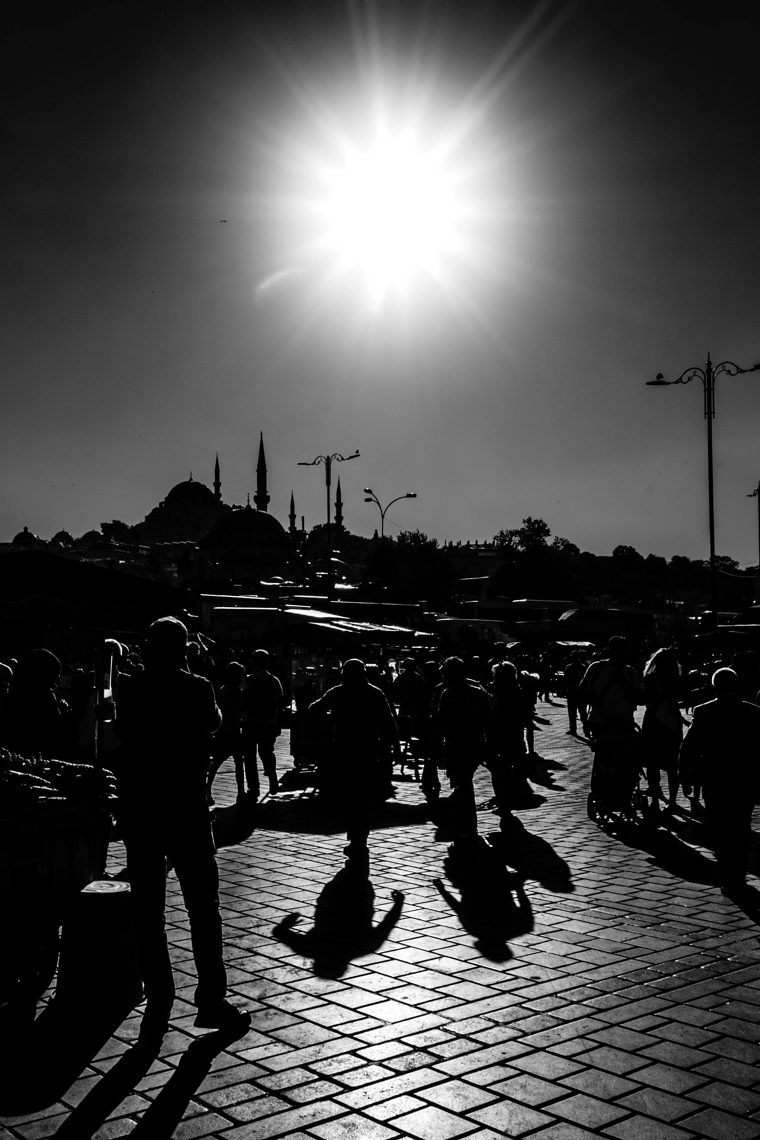 Cena de uma praça em Istambul, Turquia, com várias pessoas caminhando ao pôr do sol, incluindo uma criança em um carrinho de skateboard, sob um céu claro com o sol brilhando intensamente.