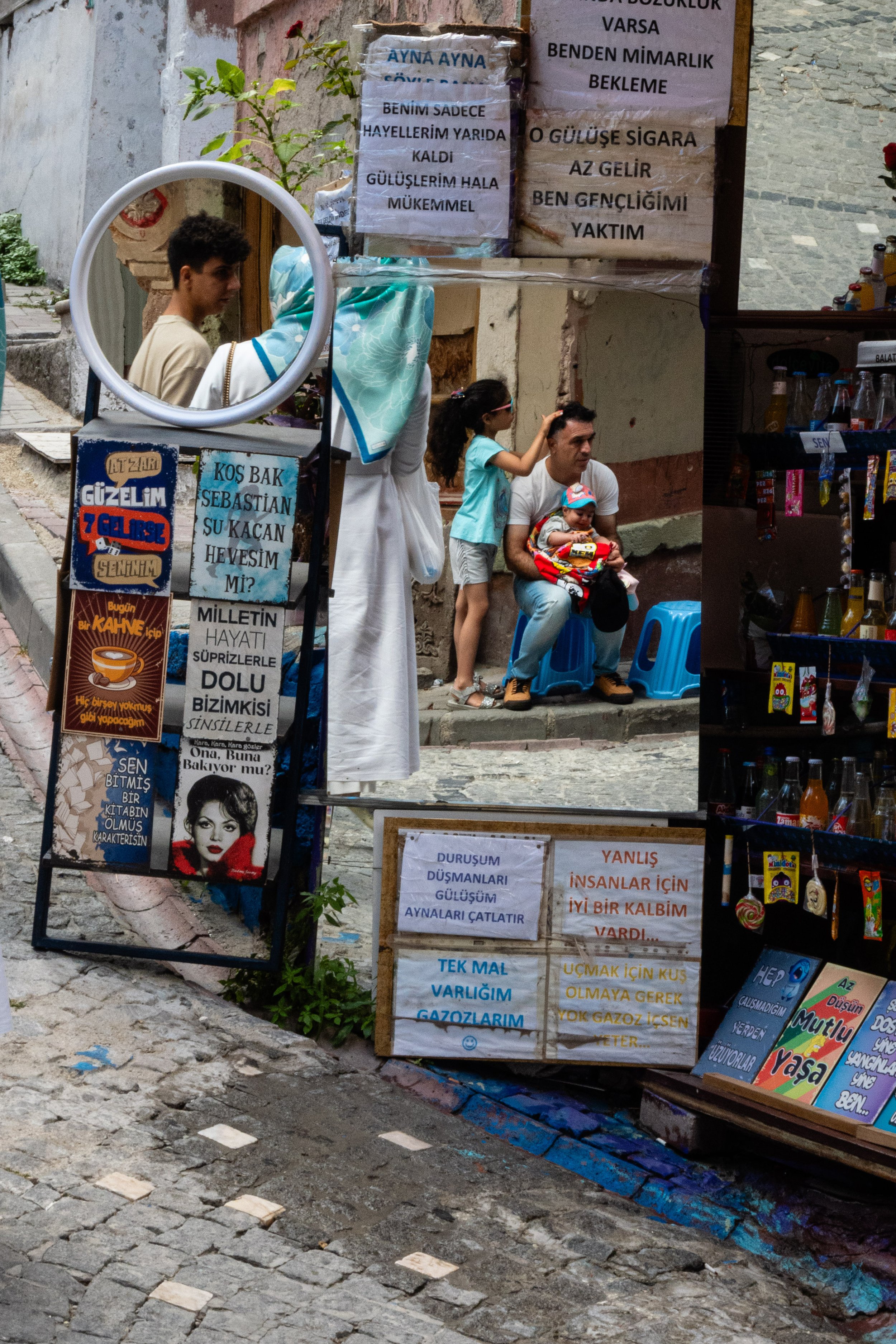 Rua de paralelepípedes com uma loja de conveniência na esquina. Vários cartazes e sinais escritos em turco, incluindo uma mensagem de texto emocional, anúncios de revendedores de bebidas e uma foto de uma mulher famosa. Pessoas na rua, incluindo um h