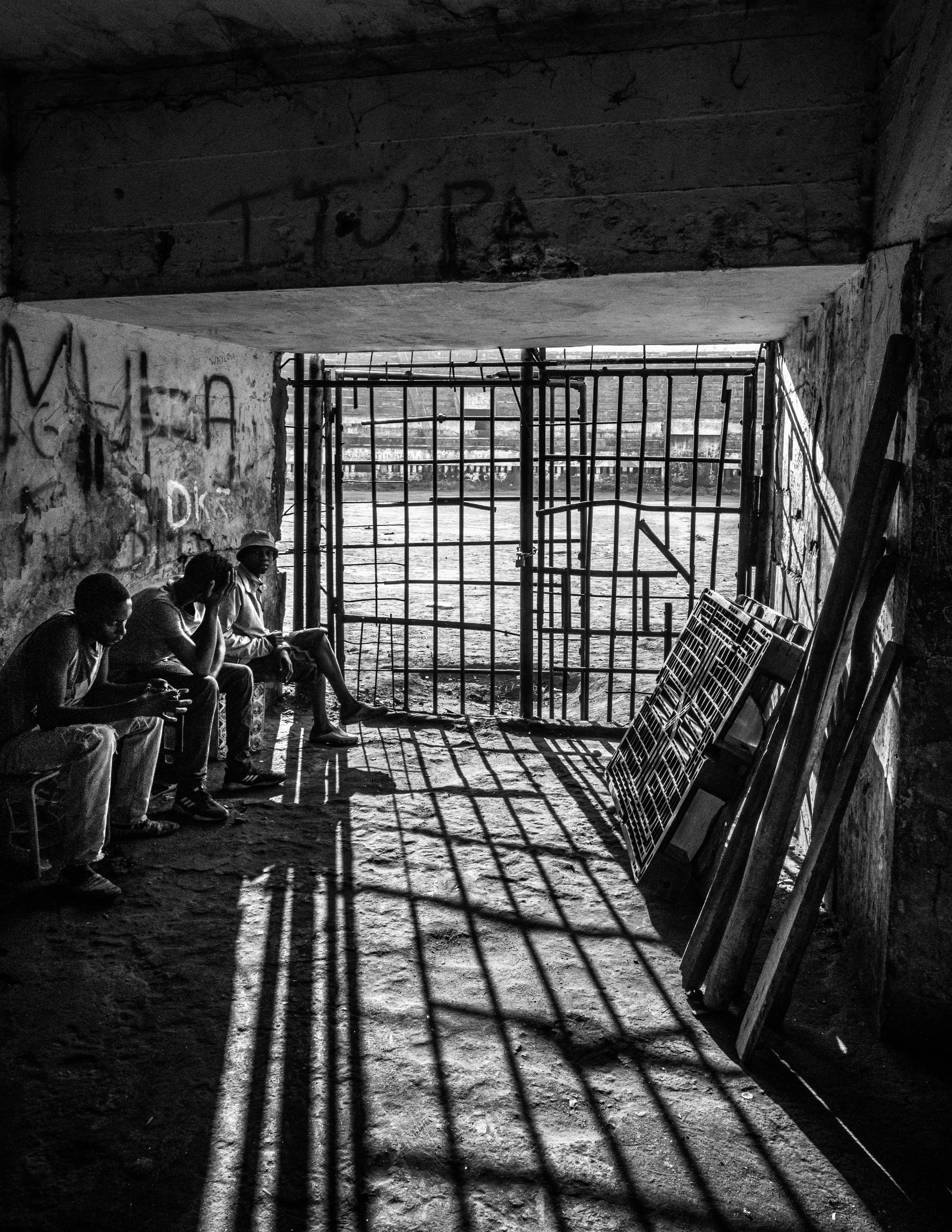 Quatro homens sentados em uma sombra de uma estrutura de ferro, com uma porta de grade atrás deles e sombras projetadas no chão de uma cela ou espaço fechado