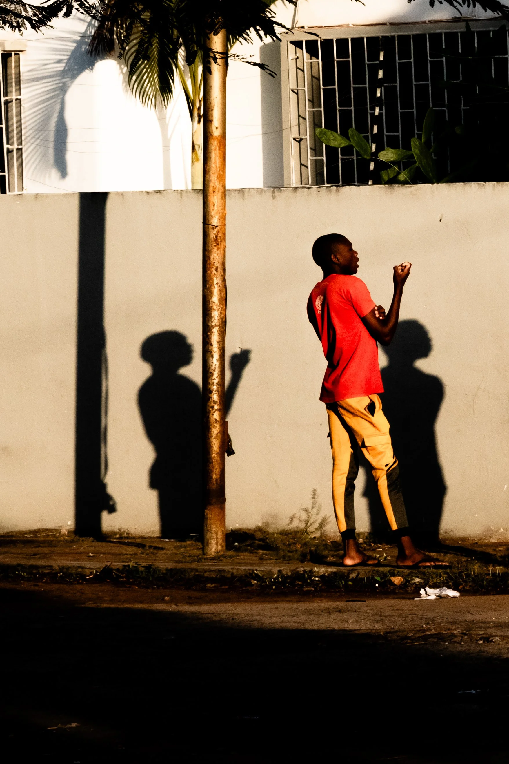 Rapaz de camiseta vermelha e calça amarela está de pé na calçada, com uma parede ao fundo, sombra dele e de uma árvore projetadas na parede, ao entardecer ou no início da noite.