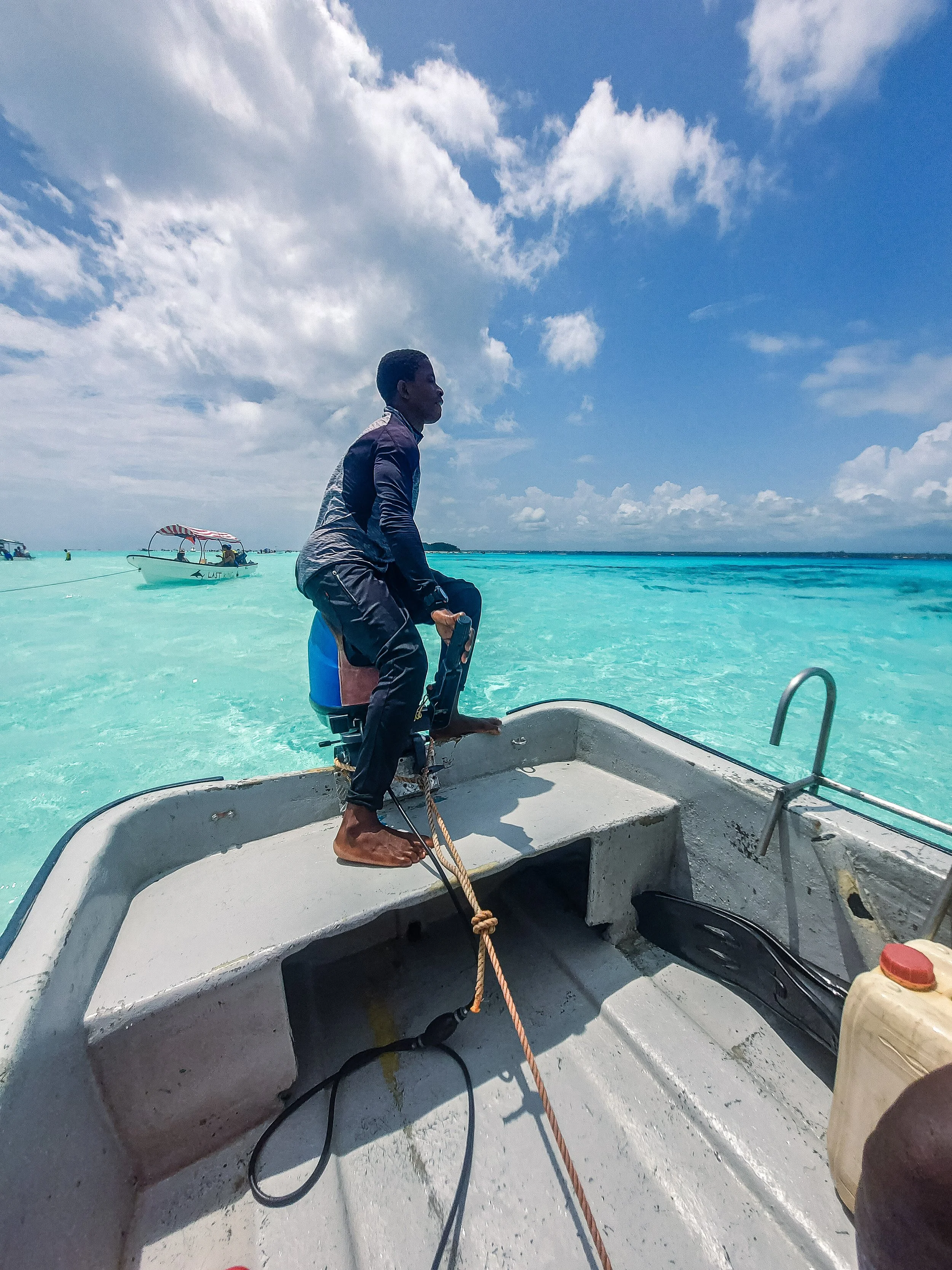 Homem sentado na proa de um barco no mar azul, com céu parcialmente nublado ao fundo.