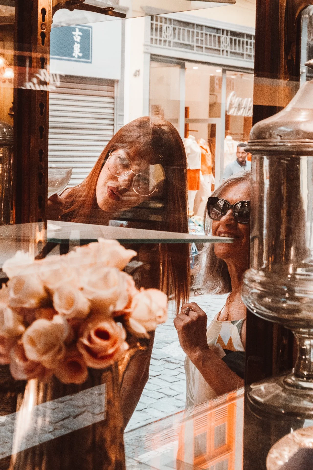 Duas mulheres observando pelo vidro de uma loja, uma usando óculos grandes e a outra óculos de grau, enquanto apreciam um vaso de flores. Ambiente urbano com lojas ao fundo.