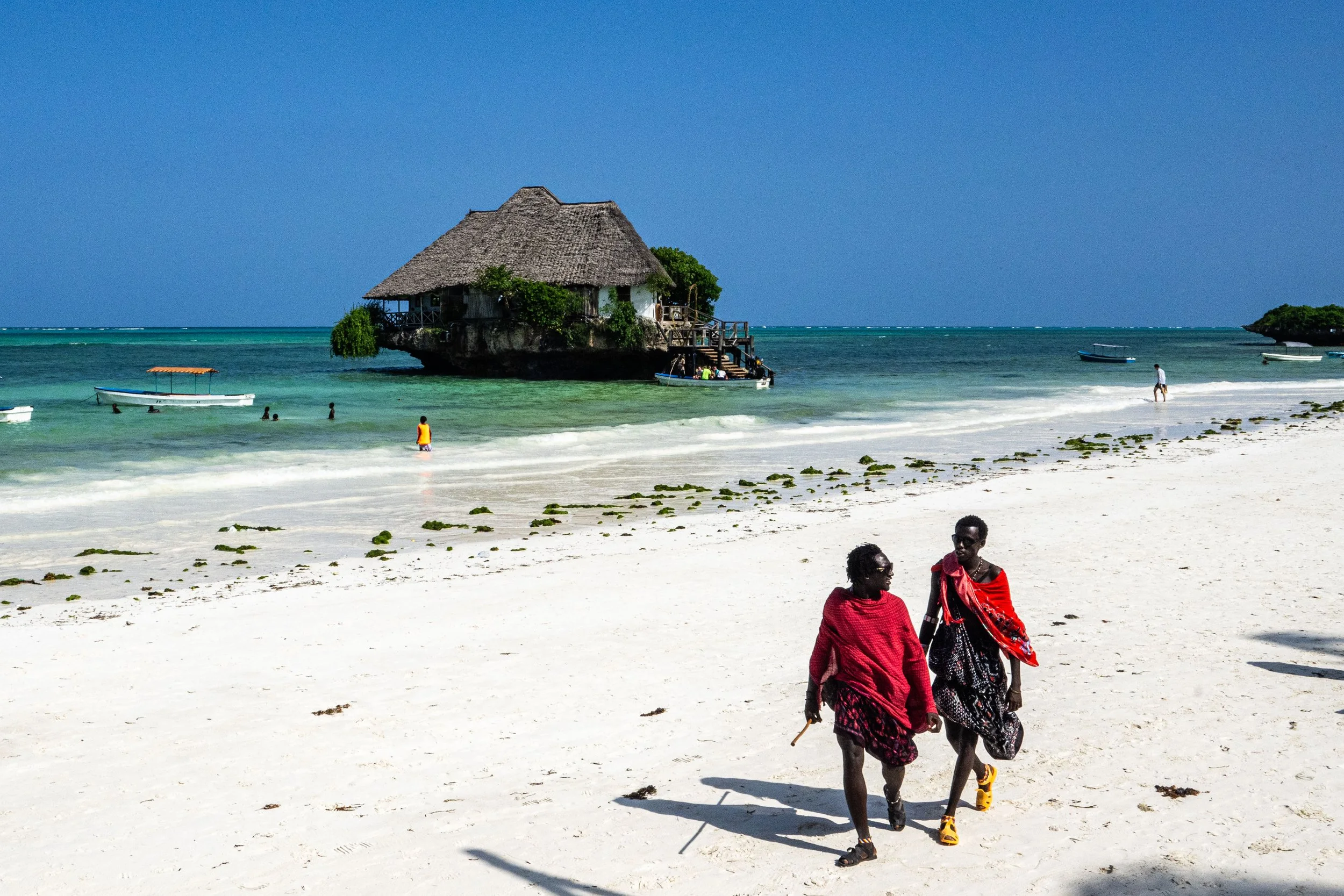 Duas pessoas vestidas com roupas vermelhas caminhando na areia de uma praia com mar azul ao fundo e uma casa de palha construída sobre uma rocha na água.
