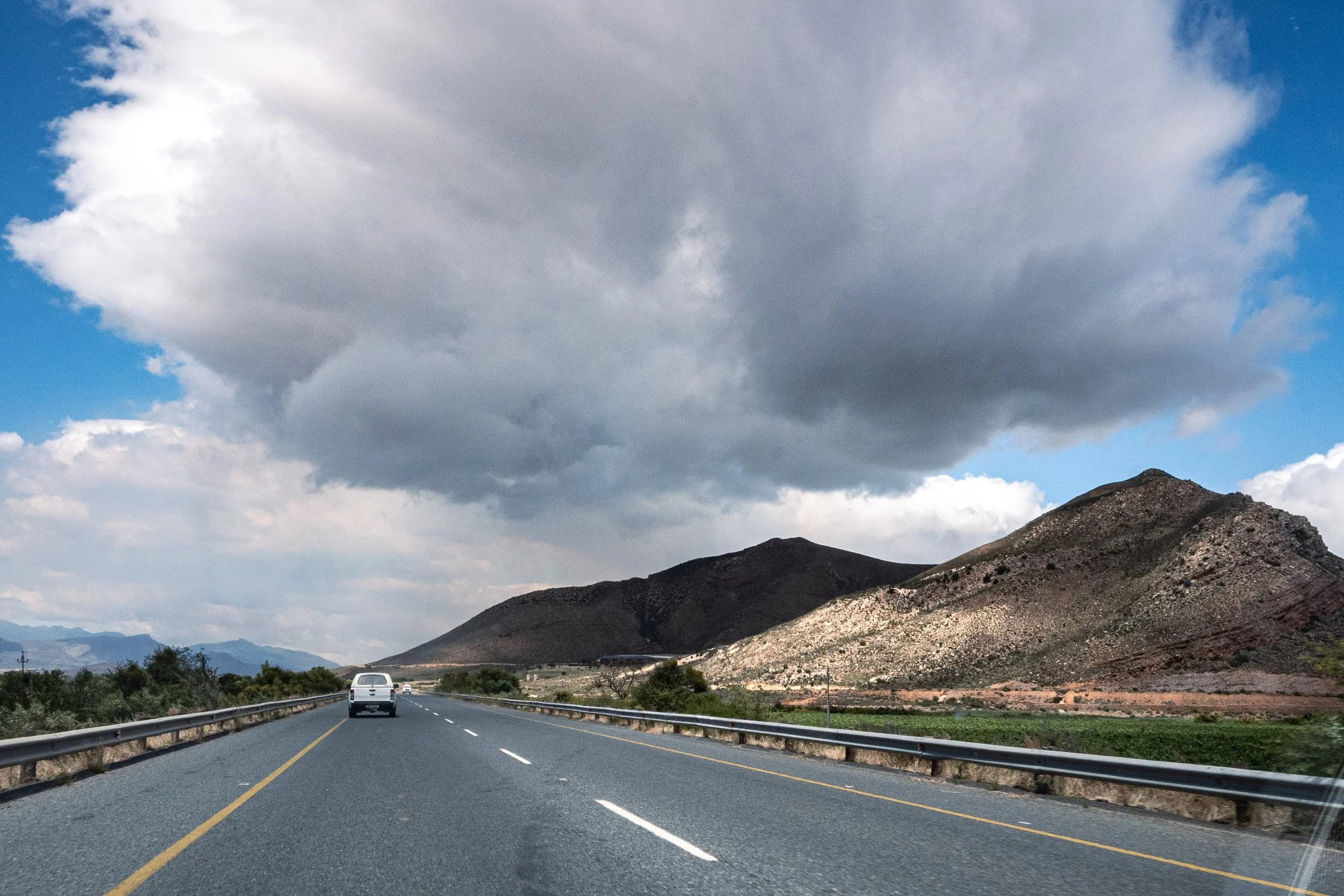 Estrada de duas faixas com carro à frente, cercada por montanhas e céu com nuvens escuras e parcialmente claras.