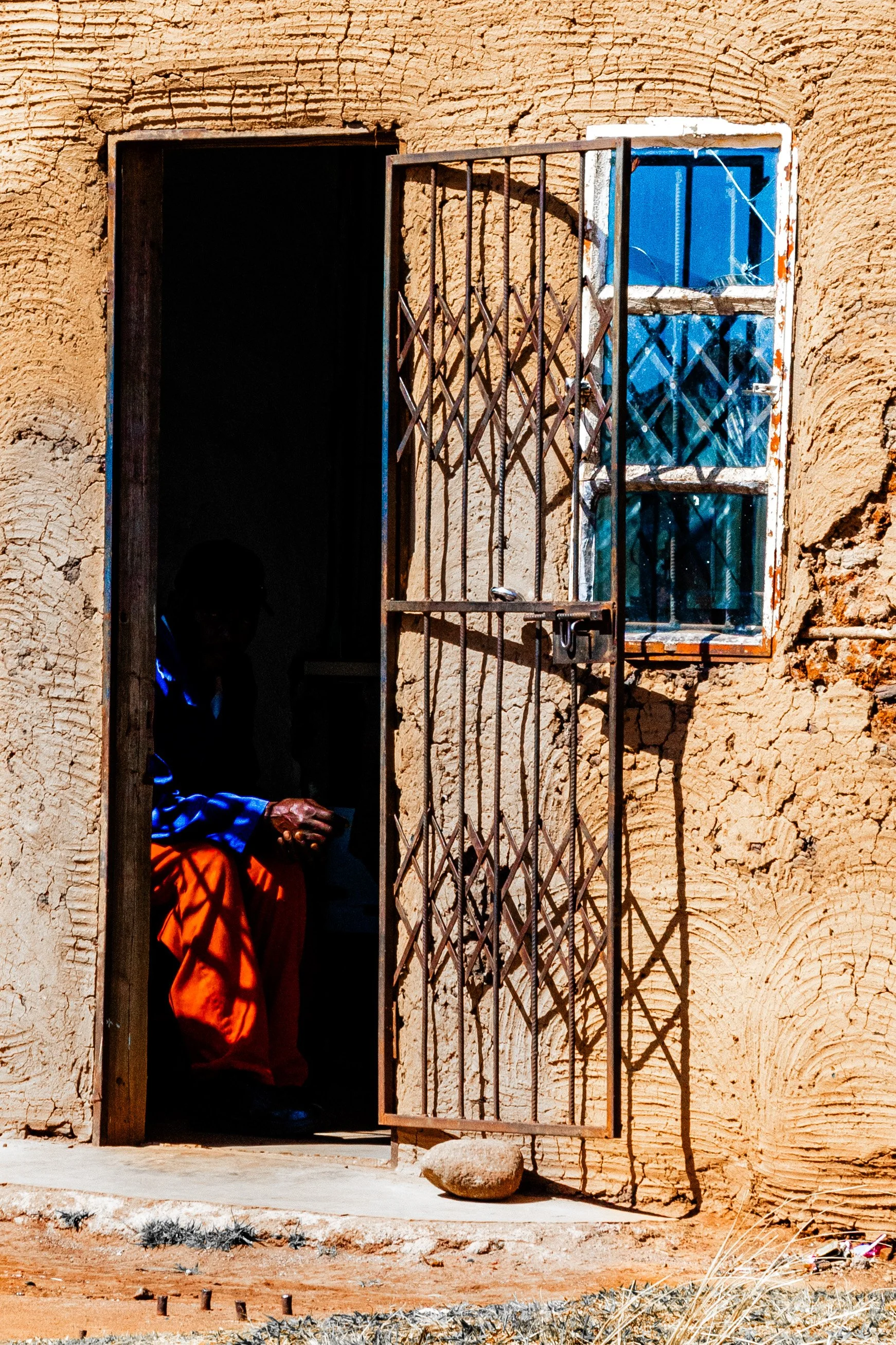 Pessoa sentada na porta de uma casa de parede de barro, com a porta aberta e uma janela com grades ao lado.