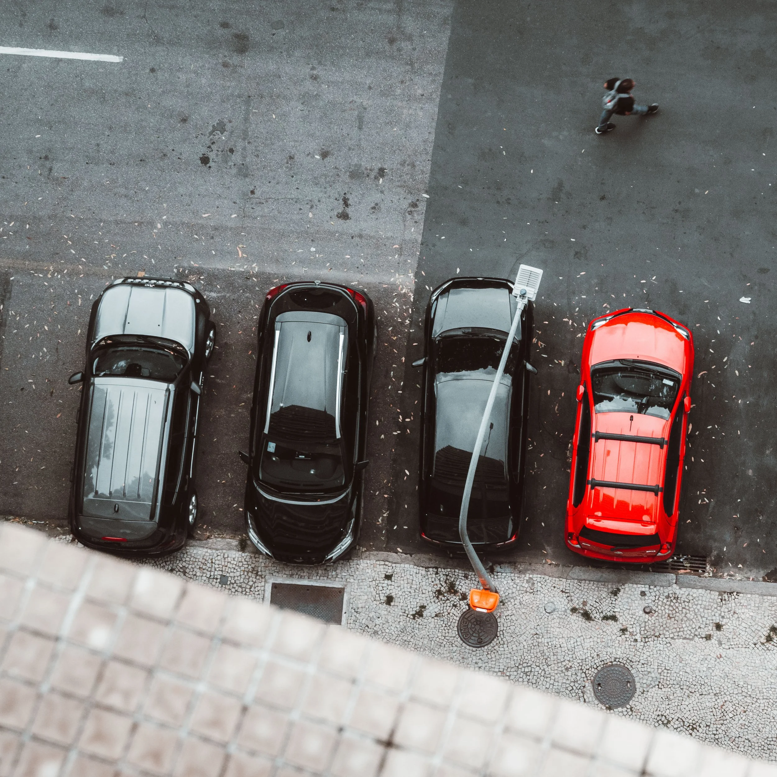 Quatro carros estacionados na rua, um deles vermelho, ao lado de um calçadão de pedras, com uma lâmpada de rua iluminando a área e uma pessoa caminhando na calçada.