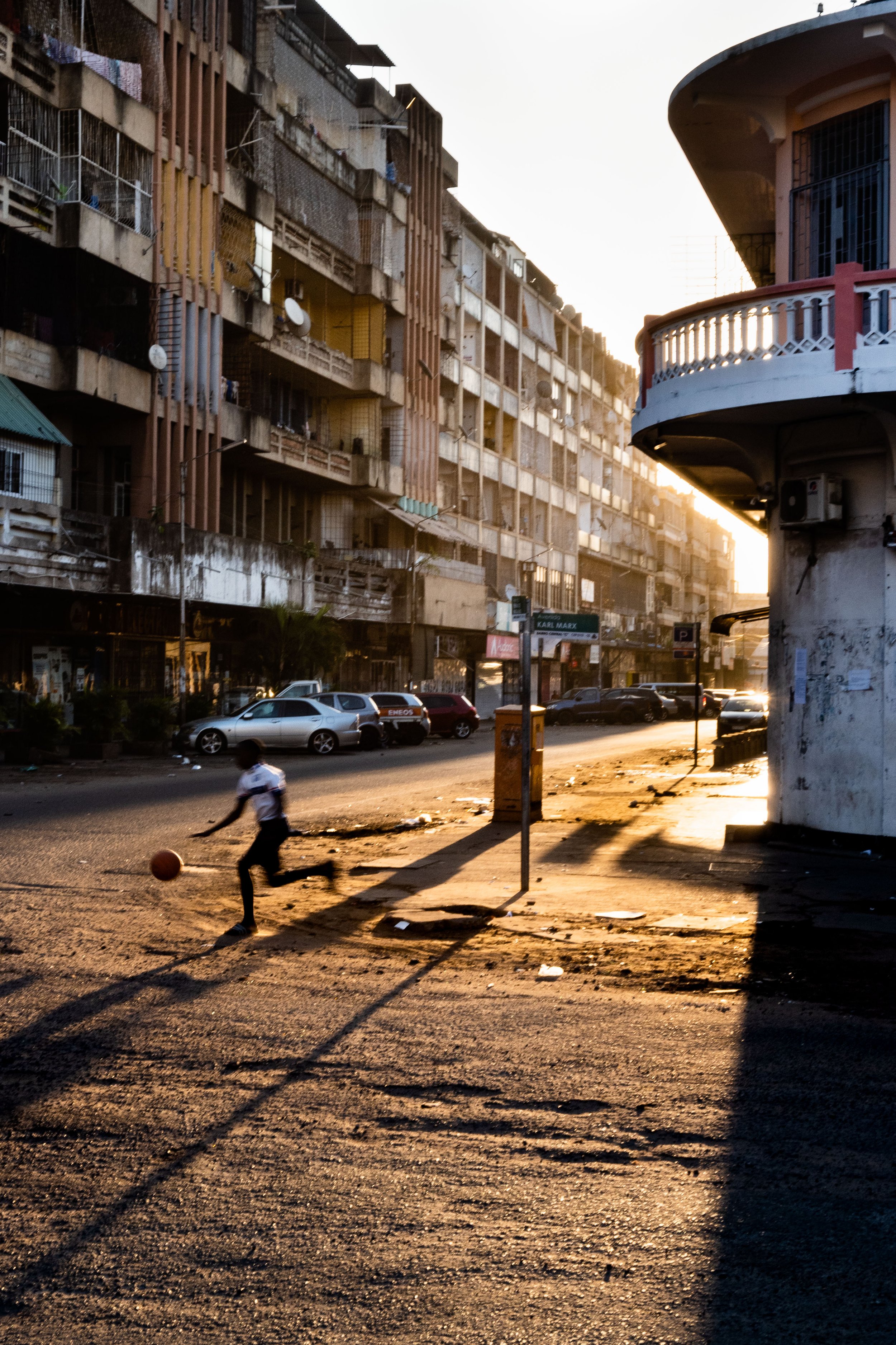 Rua de cidade com edifícios de alvenaria, carros estacionados e uma criança jogando basquete ao entardecer com forte sombra projetada ao sol.
