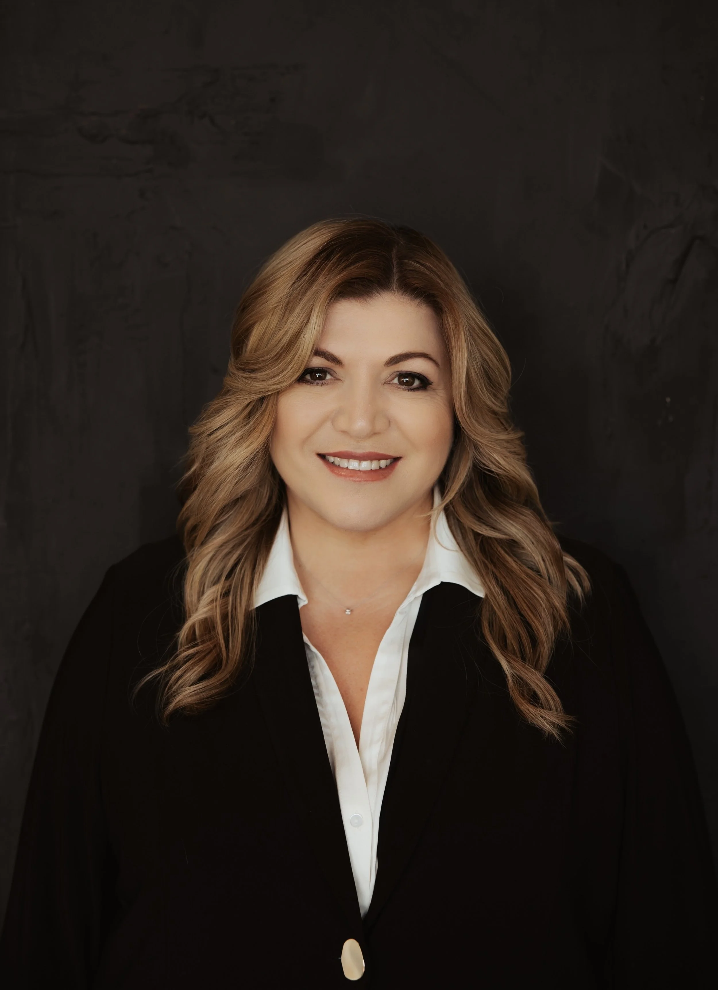 A woman with light brown, wavy hair, smiling, wearing a black blazer and a white collared shirt, posing against a dark grey textured background.