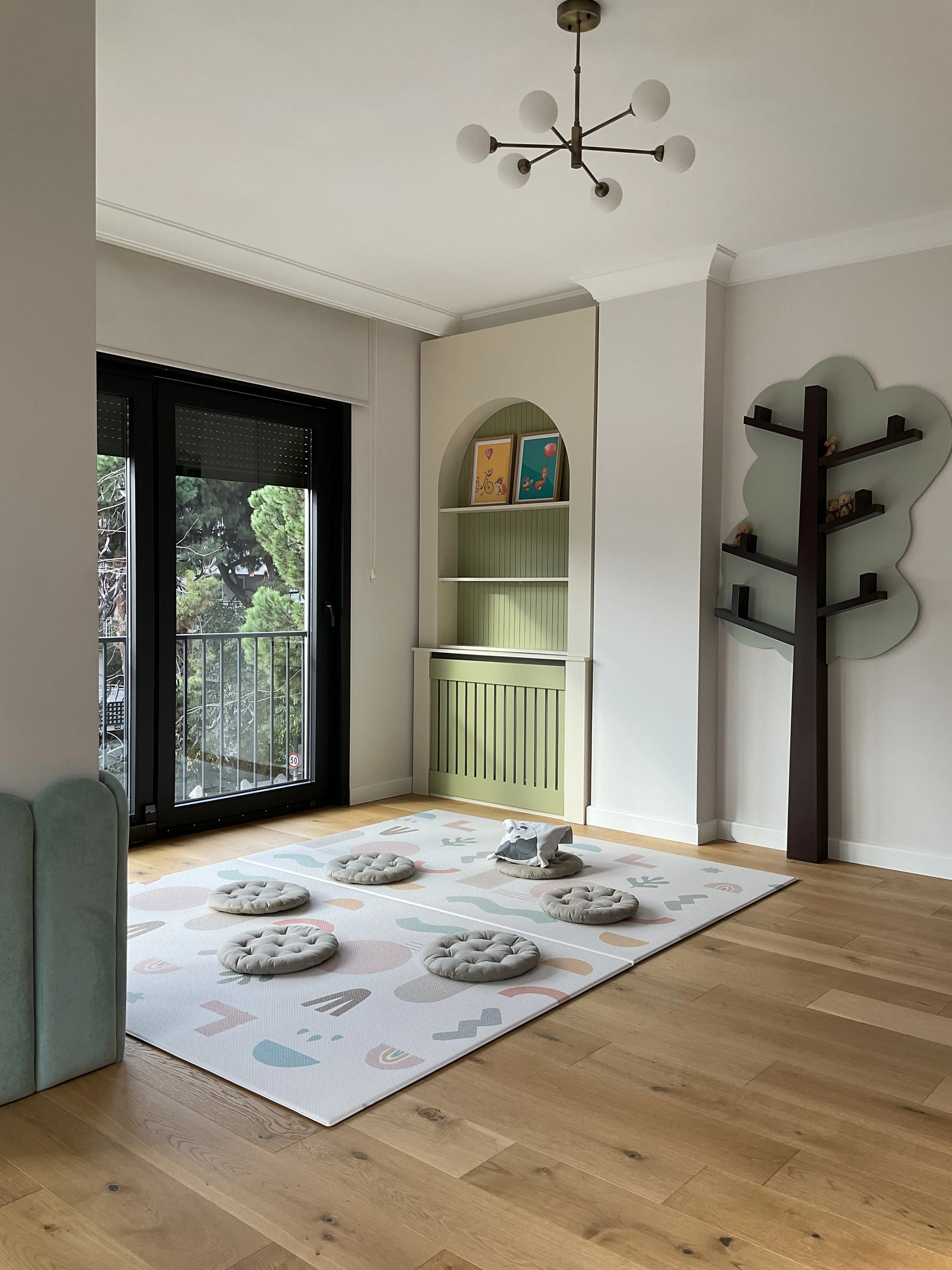 Living room with a large window, green and white built-in bookshelf, and a wall-mounted tree-shaped shelf. A white rug with pastel shapes and six beige cushions are on a wooden floor, and a small stuffed animal is on one cushion.