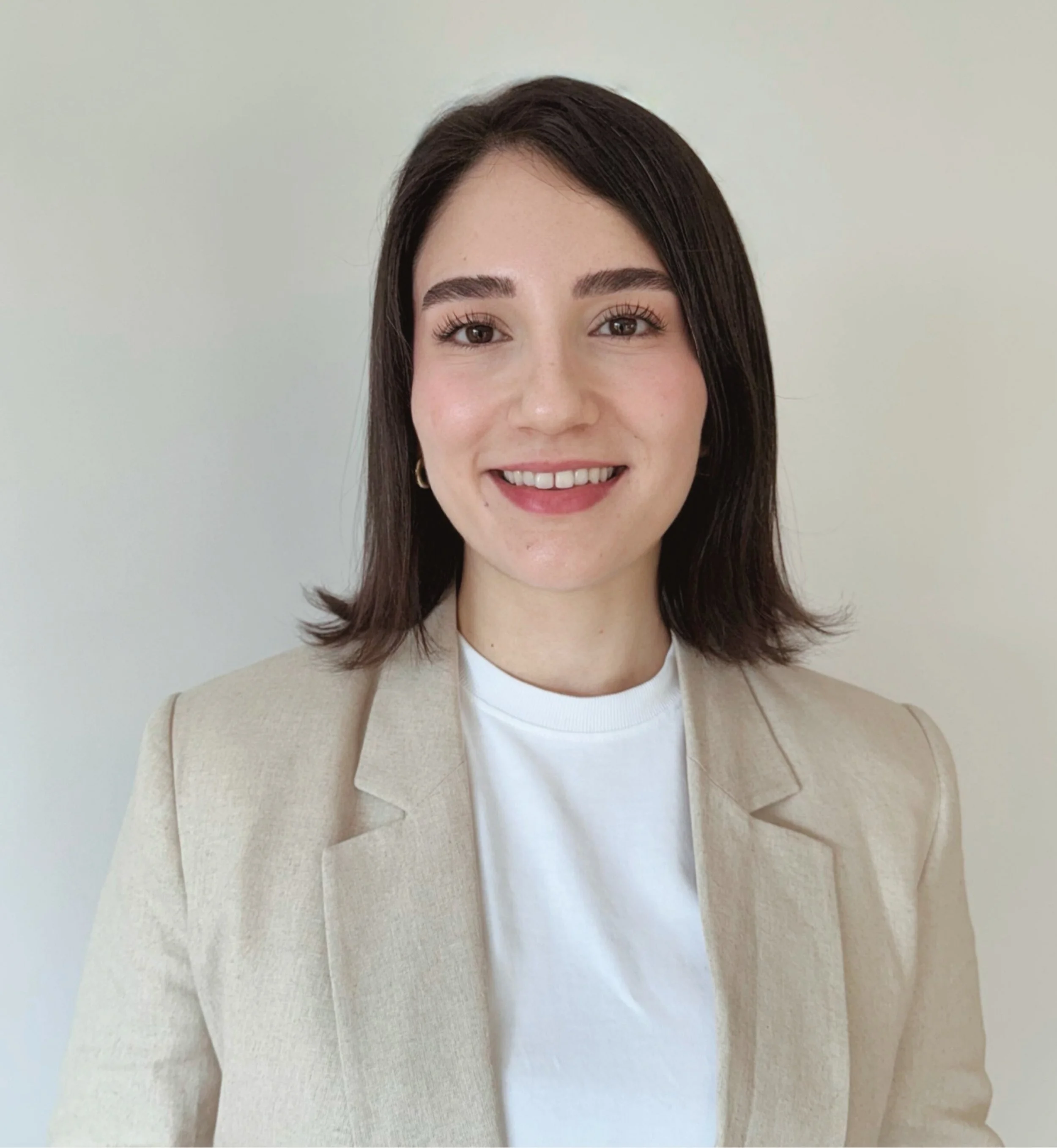 A woman with shoulder-length dark hair, wearing a beige blazer and white top, smiling against a plain light background.