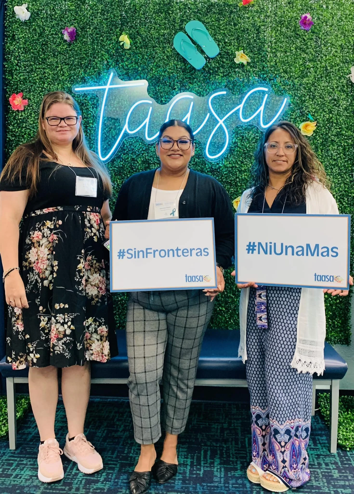 Three women standing in front of a green hedge wall with a neon sign that says "Taasa" and colorful paper flowers. They are holding signs with hashtags #SinFronteras and #NiUnaMas, and they are at an event or conference.