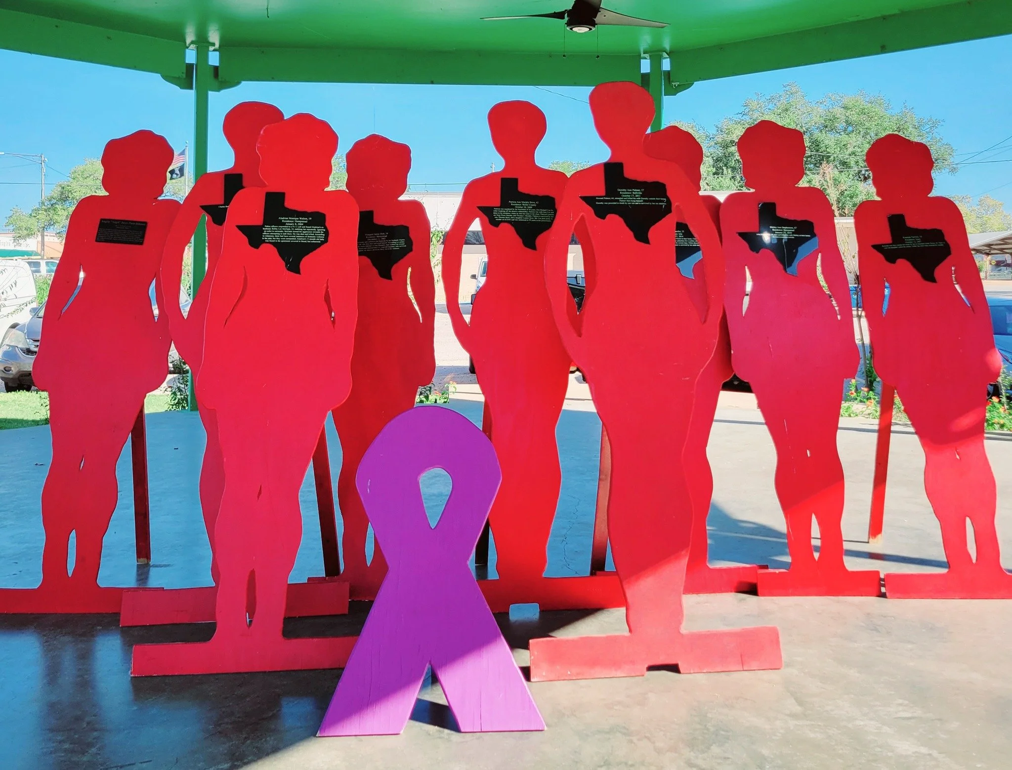 Pink and purple human-shaped cutouts with Texas-shaped cutouts, some containing text, arranged outdoors under a green roof.