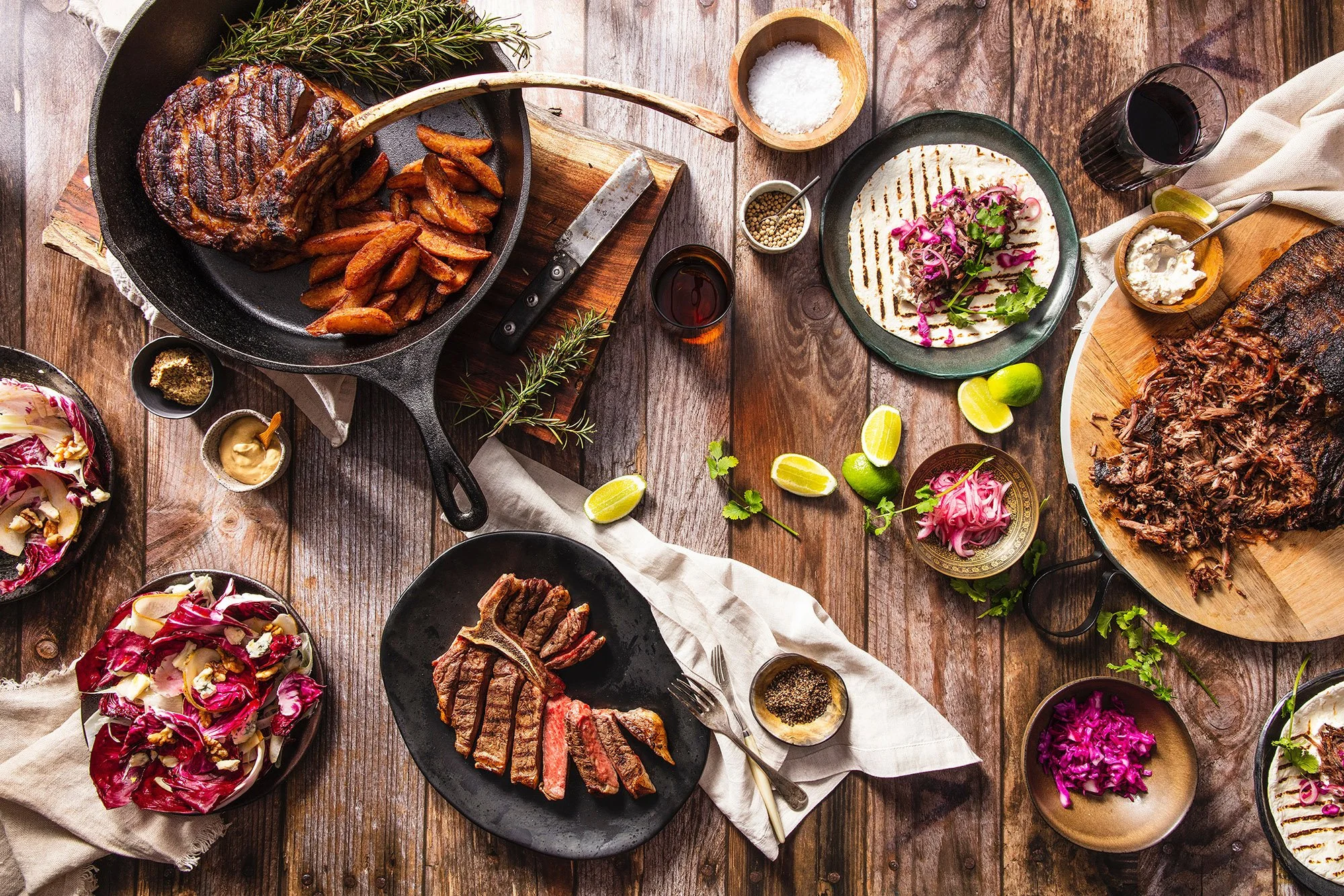 A rustic wooden table set with grilled steaks, roasted potatoes, shredded beef, colorful salads, lime wedges, and glasses of red wine, with condiments and herbs.