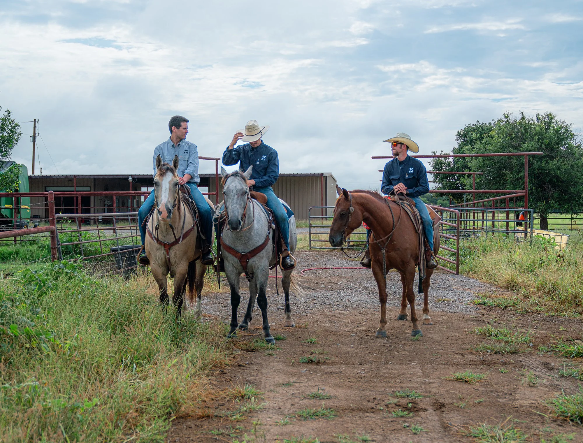 Three men on horseback in a rural farm setting with a metal barn and trees, wearing cowboy hats and casual Western attire.